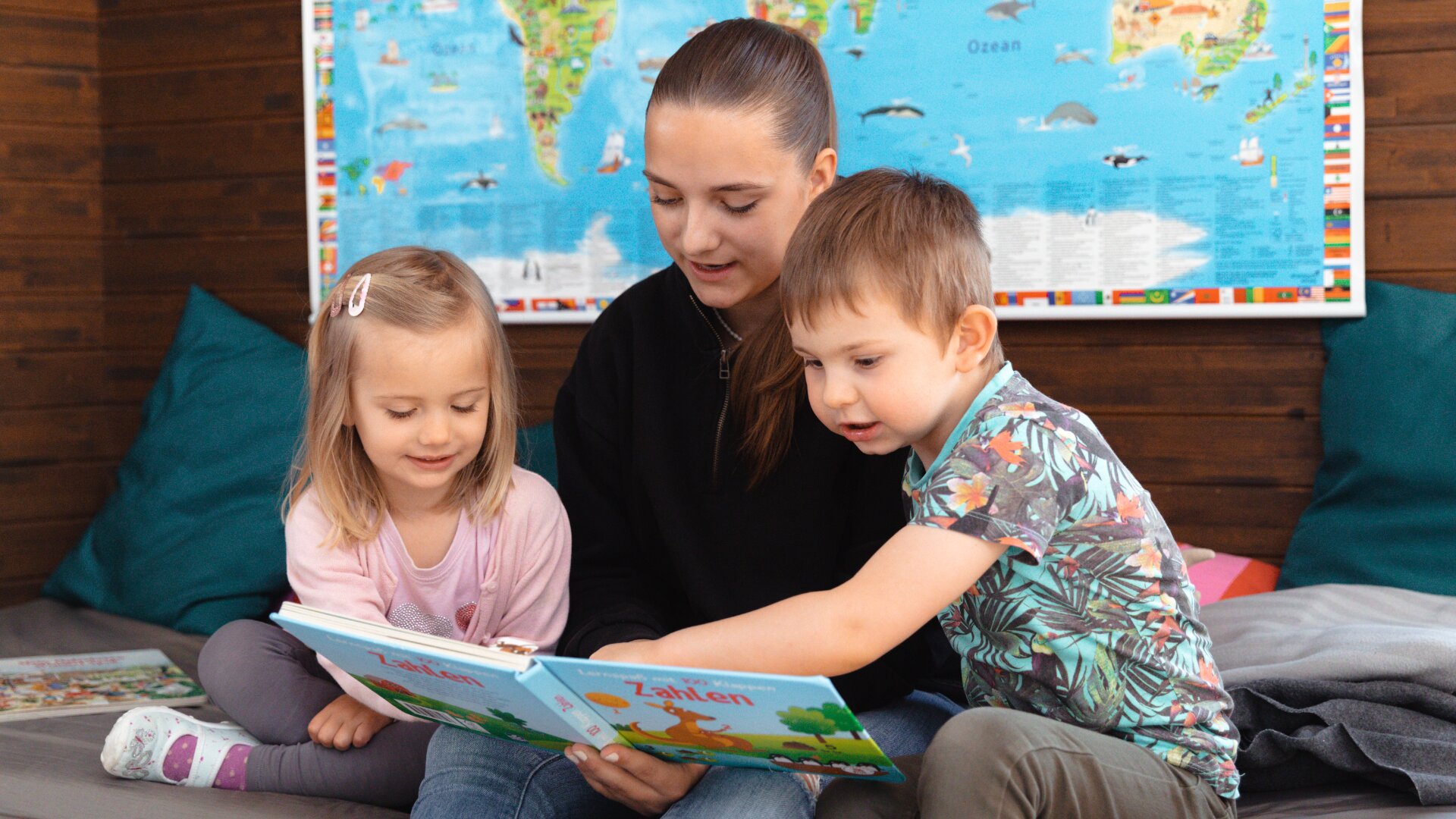 Eine Frau sitzt zwischen einem Mädchen und einem Jungen, die gemeinsam ein Buch lesen. Die Kinder sind beschäftigt und lächeln. Eine Weltkarte hängt an der Holzwand hinter ihnen.