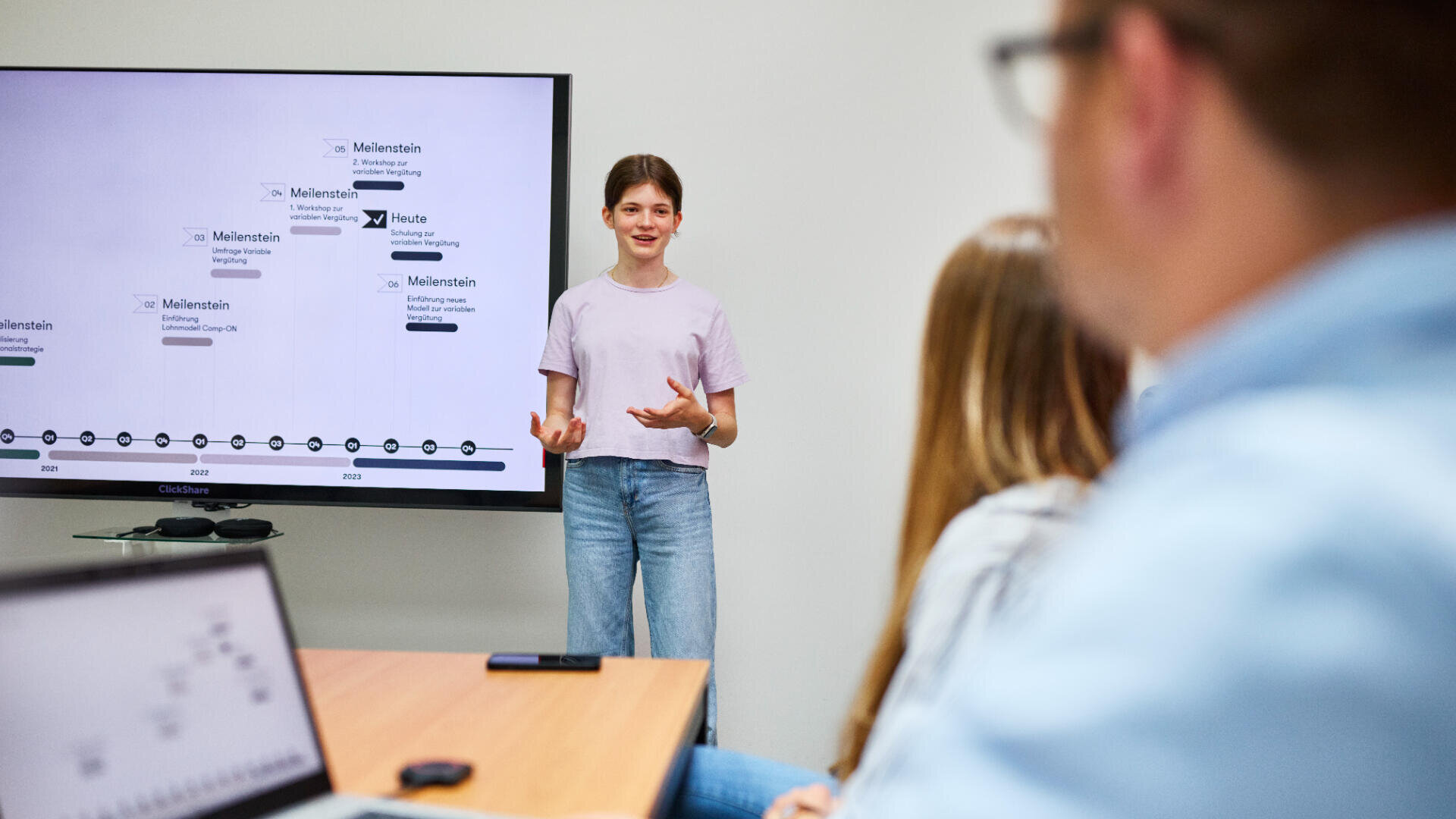 Eine junge Frau steht vor einem Bildschirm und hält eine Präsentation vor zwei sitzenden Kollegen. Auf dem Bildschirm wird eine Zeitleiste mit Text und Logos angezeigt. Auf dem Tisch im Vordergrund befinden sich Laptops und ein Smartphone.