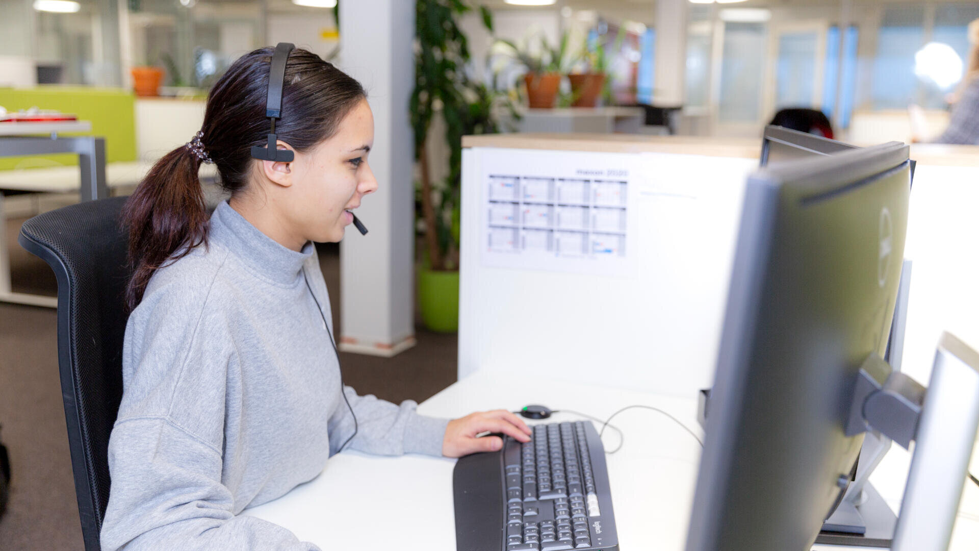 Eine Frau mit einem Headset sitzt an einem Schreibtisch in einem Büro, schaut auf einen Computermonitor und tippt auf einer Tastatur. Im Hintergrund sind Pflanzen und Büromöbel zu sehen.