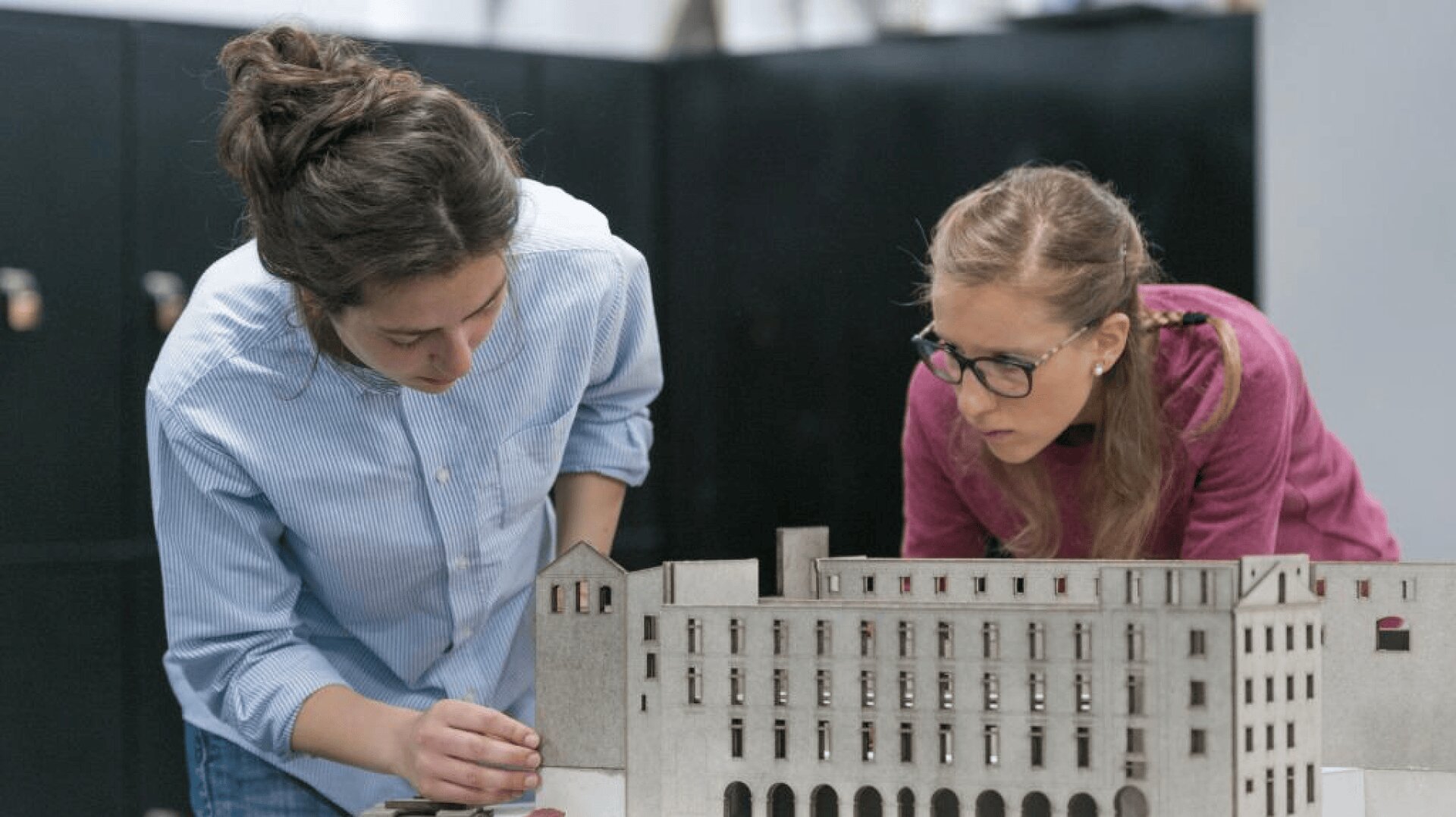 Zwei Frauen betrachten und bearbeiten ein detailliertes Architekturmodell eines großen Gebäudes. Die eine passt einen Teil des Modells an, während die andere es aufmerksam beobachtet.