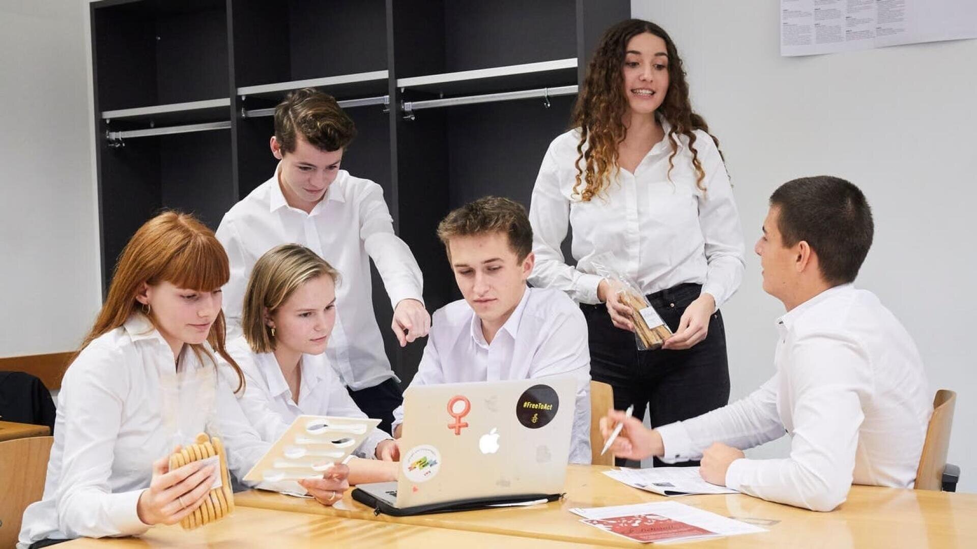 Fünf junge Erwachsene in weißen Hemden sitzen und stehen um einen Tisch mit einem Laptop, Papieren und Spielen. Zwei sitzen und diskutieren, einer zeigt auf den Laptop, ein anderer steht und spricht und hält Holzklötze.