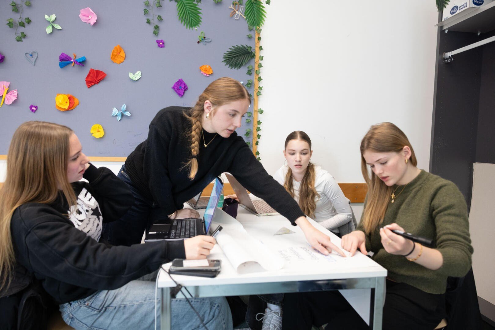 Vier junge Frauen sitzen an einem Tisch und arbeiten mit Papieren und Laptops an einem Gruppenprojekt. Eine Frau deutet auf ein Blatt, während die anderen sich konzentrieren. Im Hintergrund ist eine dekorierte Pinnwand mit Blumen und Blättern zu sehen.