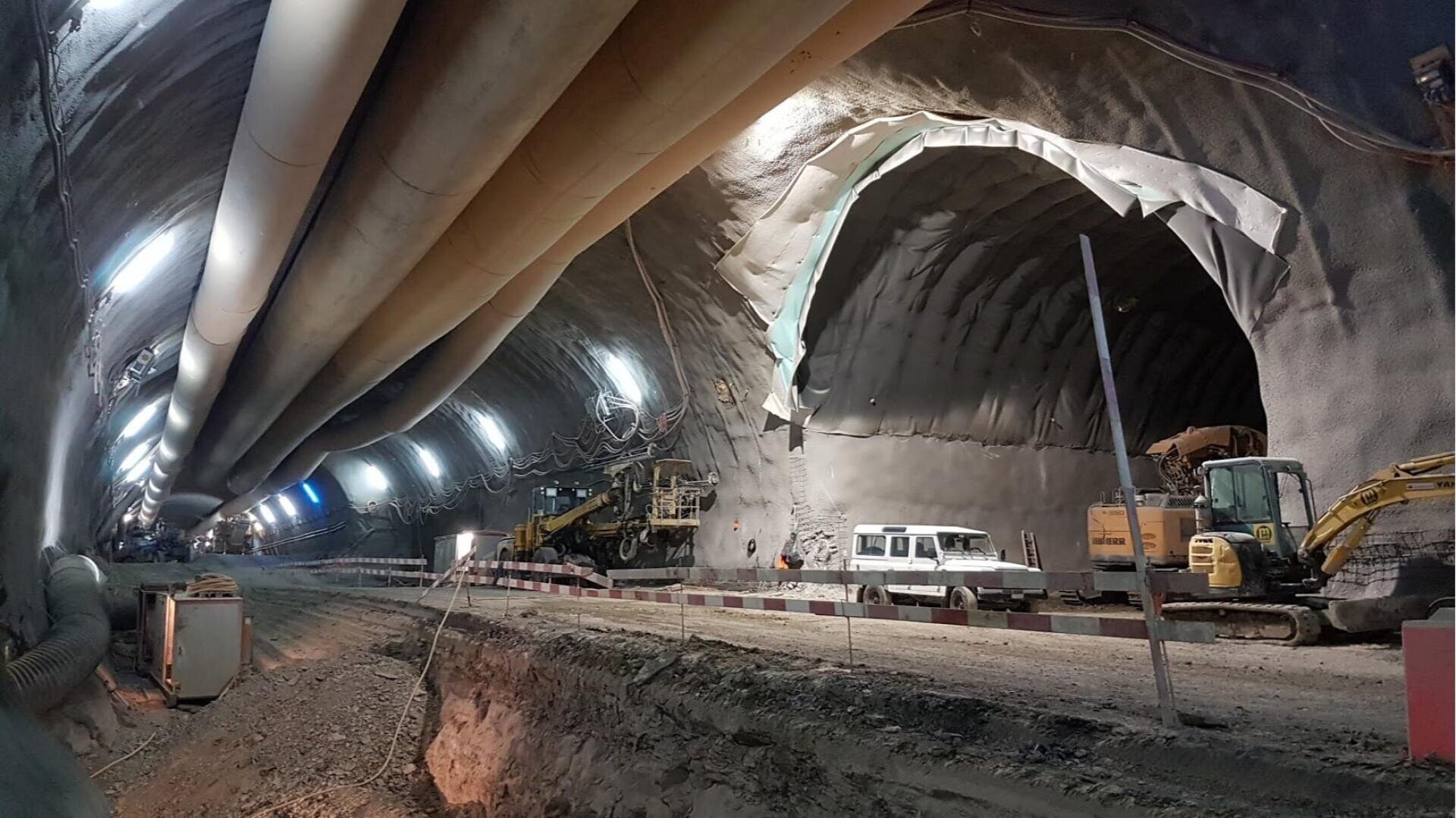 Bauarbeiten im Inneren eines großen Tunnels, mit Aushubgeräten, einem weißen Nutzfahrzeug und Oberlicht. Die Tunnelwände sind verstärkt, und an der Decke verlaufen Rohre. Auf einer Seite des Tunnels wird ein großes Loch gegraben.