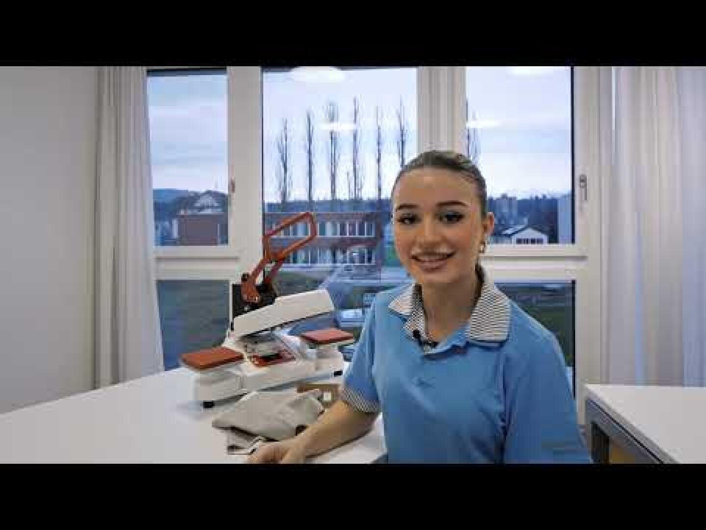 Eine junge Frau in einer blauen Uniform lächelt in die Kamera, während sie an einem Tisch mit einer Heißpressmaschine und gefaltetem Stoff sitzt. Große Fenster hinter ihr zeigen Bäume und ein Gebäude im Freien.