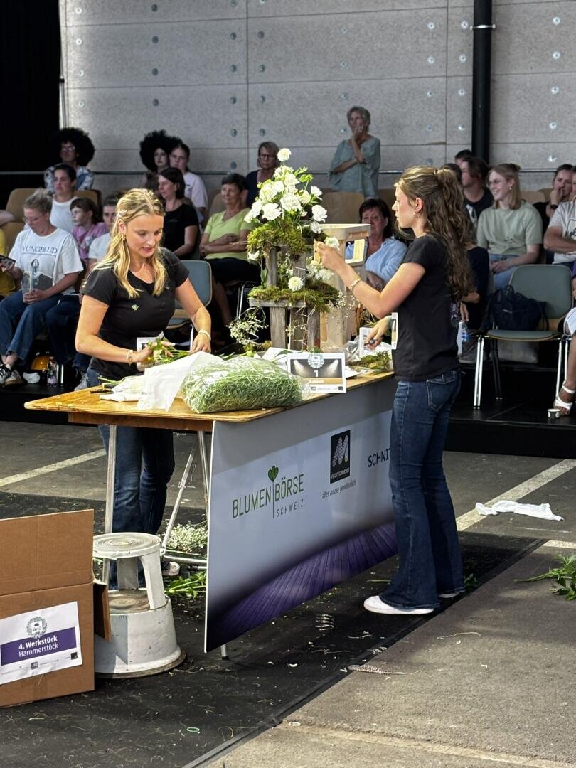 Zwei Frauen arrangieren während einer öffentlichen Veranstaltung auf der Bühne an einem Tisch Blumen, während das Publikum zusieht. Auf dem Tisch liegen Blumenutensilien, und in der Mitte steht ein weißes Blumenarrangement.