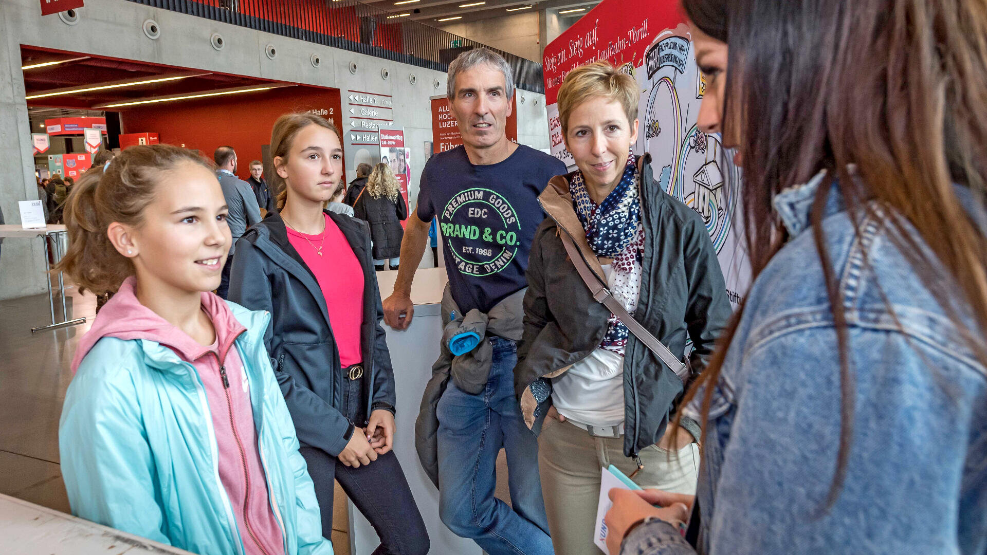 Eine Gruppe von fünf Personen, darunter zwei Mädchen, ein Mann und zwei Frauen, steht in einem Innenraum und unterhält sich. Sie sind leger gekleidet und scheinen auf einer öffentlichen Veranstaltung oder Ausstellung zu sein.