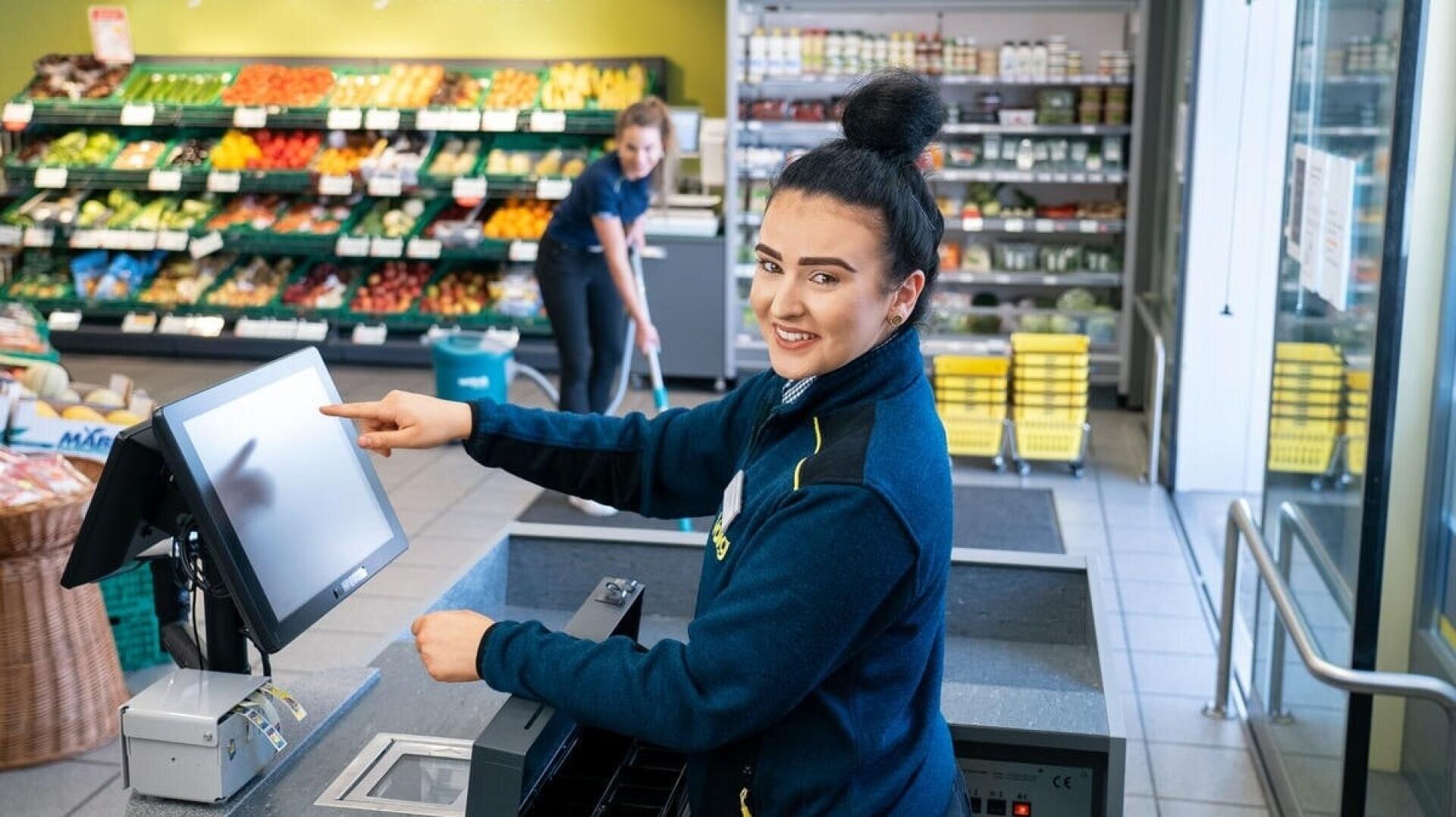 Eine Supermarktkassiererin lächelt und zeigt auf einen Touchscreen an der Kasse, während ein anderer Mitarbeiter im Hintergrund den Boden wischt. Hinter ihnen sind Regale mit frischen Produkten zu sehen.