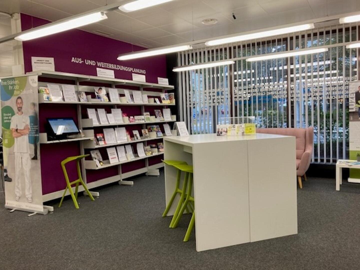 Eine moderne Bibliothek mit lilafarbenen Wänden, Bücherregalen voller Broschüren, einem weißen Tisch mit grünen Stühlen, einem rosafarbenen Sessel und großen Fenstern mit vertikalen Jalousien, die natürliches Licht hereinlassen.