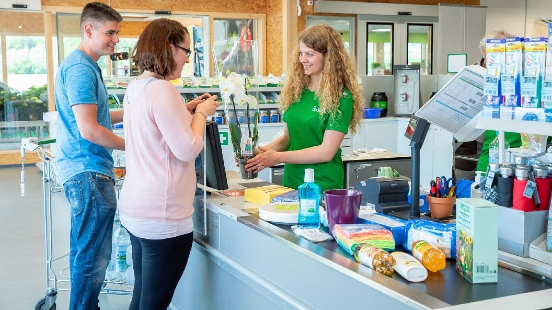 Eine Kassiererin in einem grünen Hemd lächelt eine Frau an, die an einer Kasse Lebensmittel und eine Topforchidee bezahlt, während ein Mann mit einem Einkaufswagen hinter ihr steht.