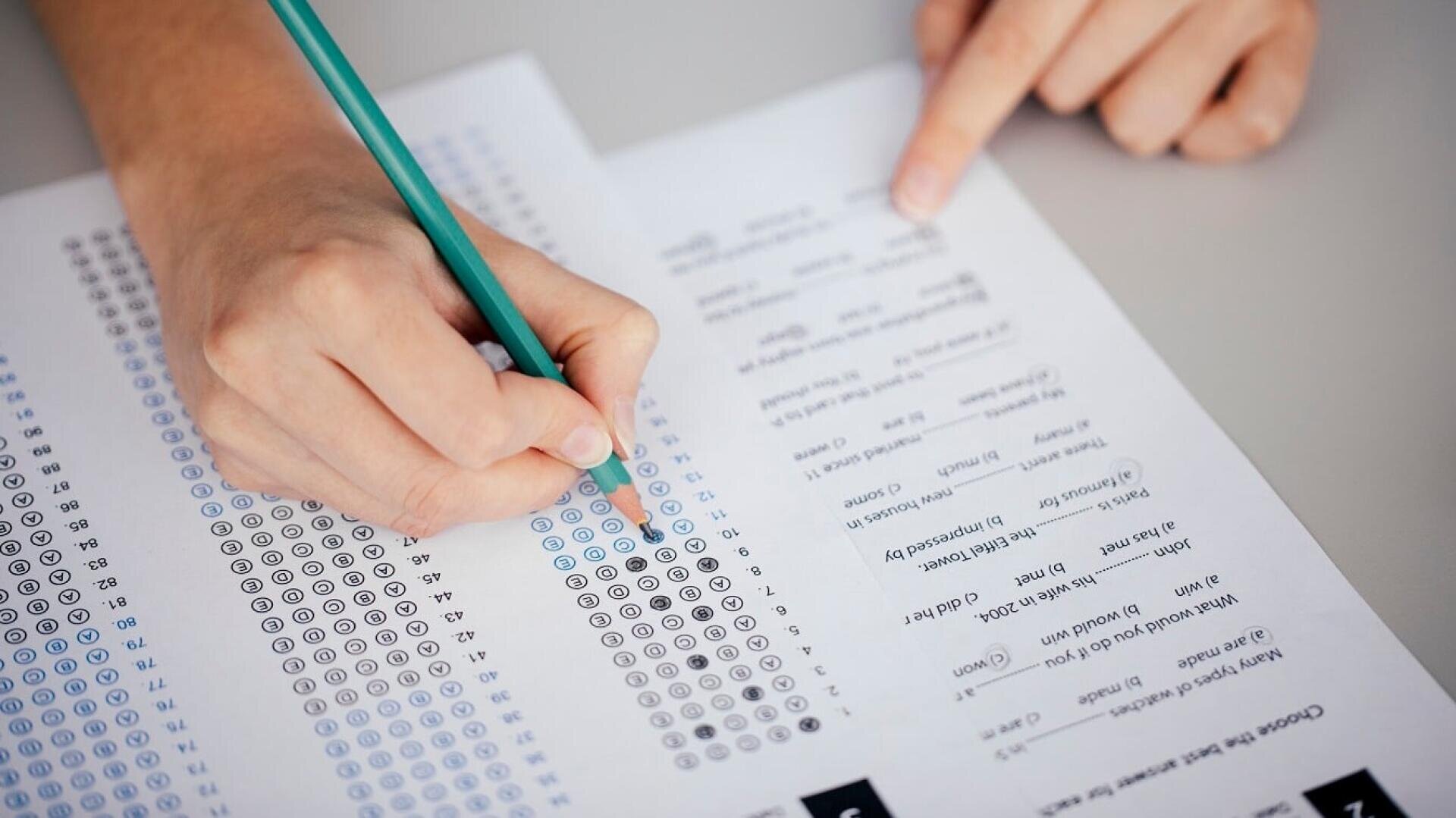 Eine Person füllt die Antwortfelder auf einem Multiple-Choice-Testbogen mit einem Bleistift aus, während sie auf eine Frage auf einem anderen Blatt Papier zeigt.