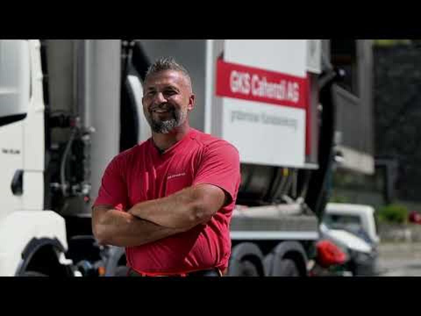 Ein Mann in einem roten Hemd steht lächelnd mit verschränkten Armen vor einem großen Lkw, auf dem ein Schild mit der Aufschrift „GIS Cabral AG“ zu sehen ist.