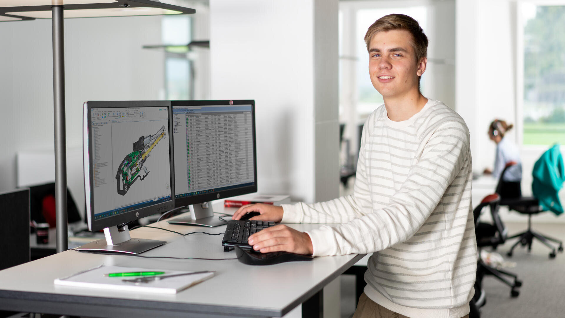 Eine junge Person in einem gestreiften Pullover steht an einem Schreibtisch und arbeitet in einem hellen, modernen Büro an einem Computer mit zwei Monitoren, auf denen Ingenieur- oder Design-Software angezeigt wird.