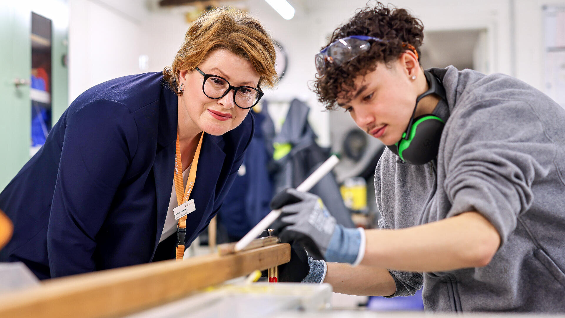 Eine Frau mit Brille beobachtet einen jungen Mann, der eine Schutzausrüstung trägt, während er in einer Werkstatt ein Holzbrett sorgfältig ausmisst und bearbeitet.