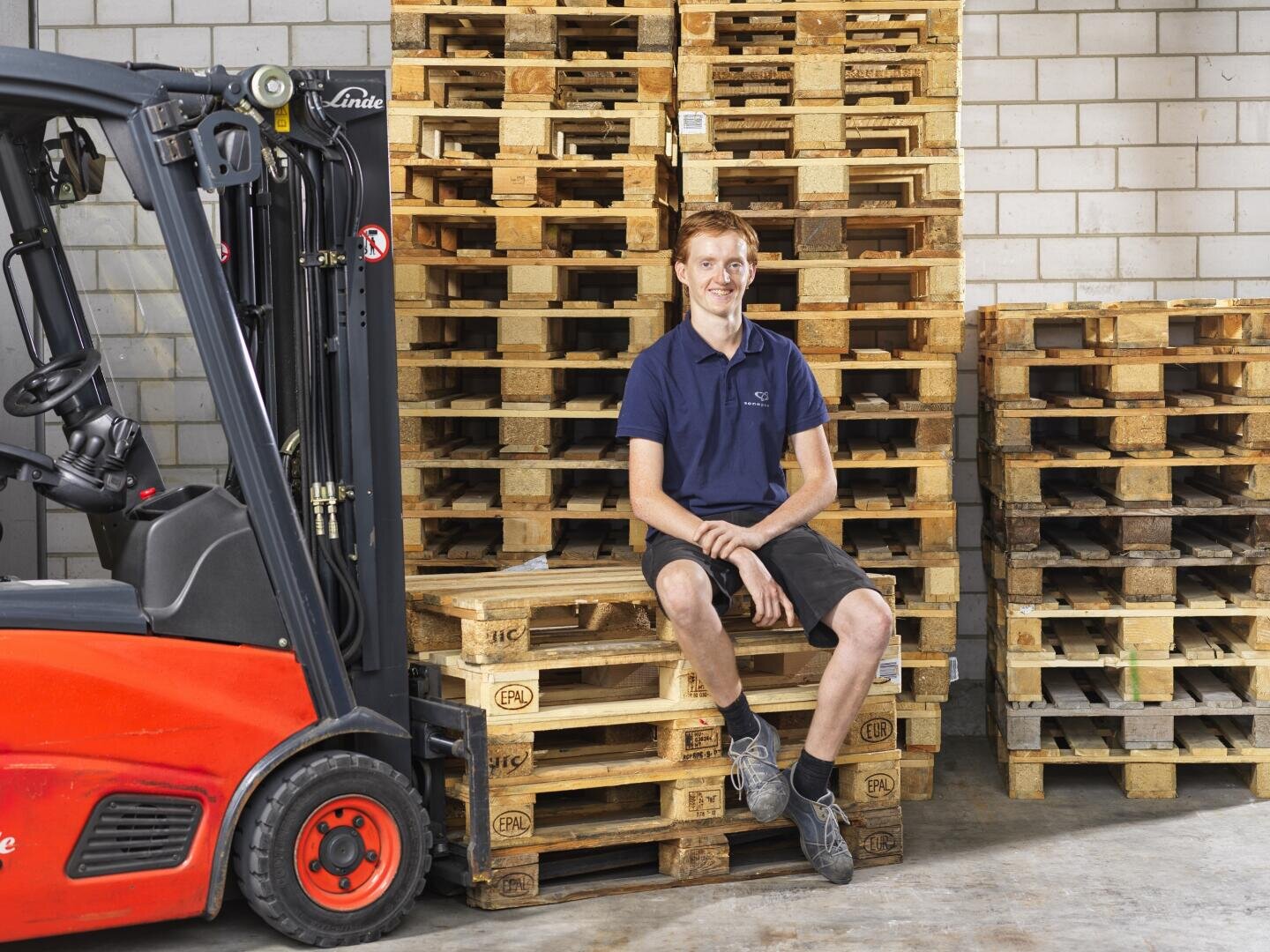 Ein junger Mann in einem marineblauen Polohemd und Shorts sitzt auf einem Stapel Holzpaletten in einem Lagerhaus und lächelt. Links ist ein roter Gabelstapler geparkt, und weitere Holzpaletten sind an einer Backsteinwand gestapelt.