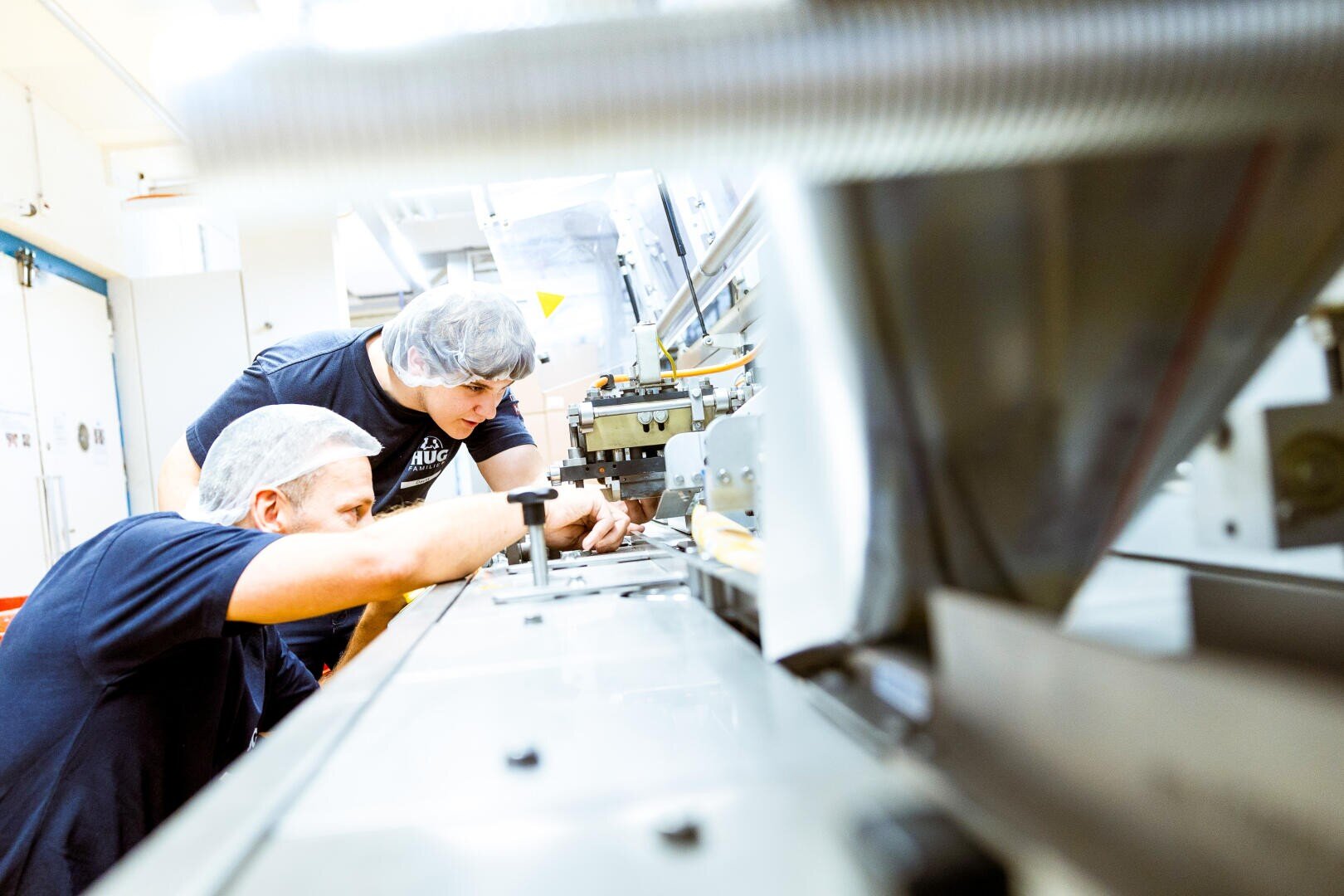 Zwei Personen, die ein Haarnetz und ein dunkles Hemd tragen, inspizieren oder arbeiten an einer großen Industriemaschine in einer hellen, sauberen Anlage.