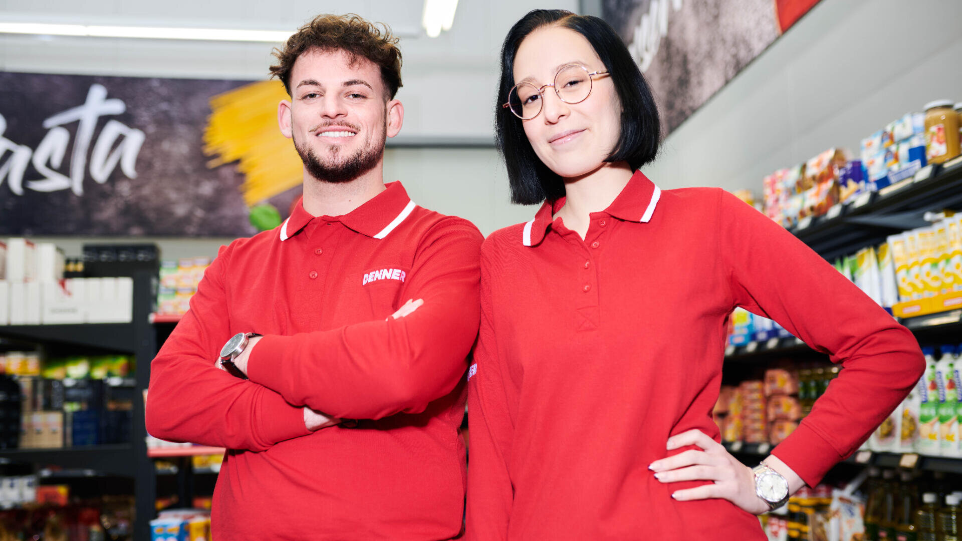 Zwei Supermarktangestellte in roten Uniformen stehen selbstbewusst in einem Gang, umgeben von Regalen mit Lebensmittelprodukten. Der eine hat die Arme verschränkt, der andere eine Hand in die Hüfte gestemmt.