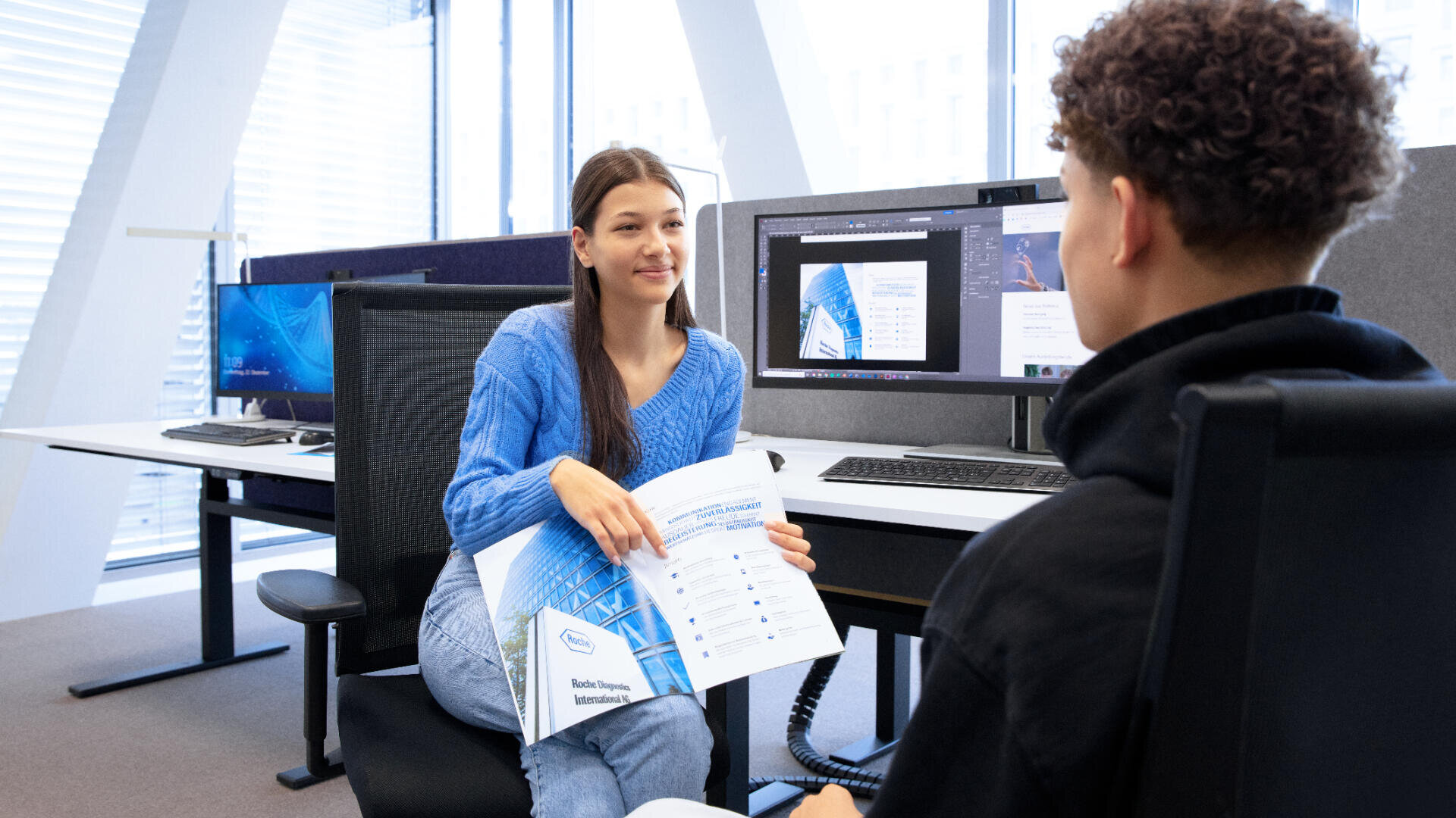 Eine Frau in einem blauen Pullover sitzt an einem Schreibtisch mit einem ausgedruckten Bericht und spricht mit einem Mann in einer Büroumgebung, in der im Hintergrund Computermonitore mit Dokumenten zu sehen sind.