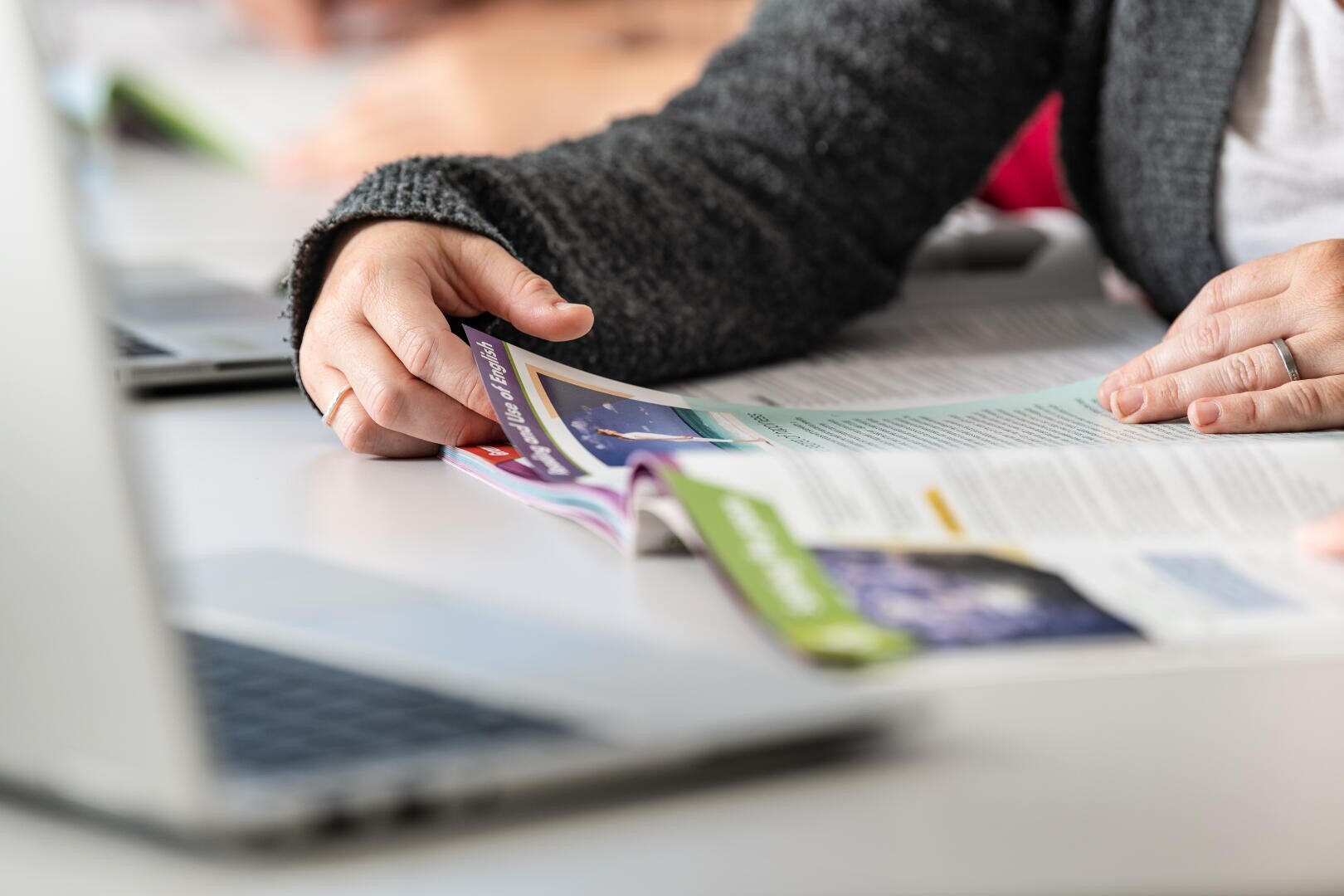 Eine Person, die einen dunklen Pullover trägt, liest an einem Schreibtisch eine bunte Zeitschrift oder Broschüre, während in der Nähe ein Laptop geöffnet ist. Die Person blättert eine Seite um, und nur ihre Hände und ein Teil ihres Arms sind sichtbar.