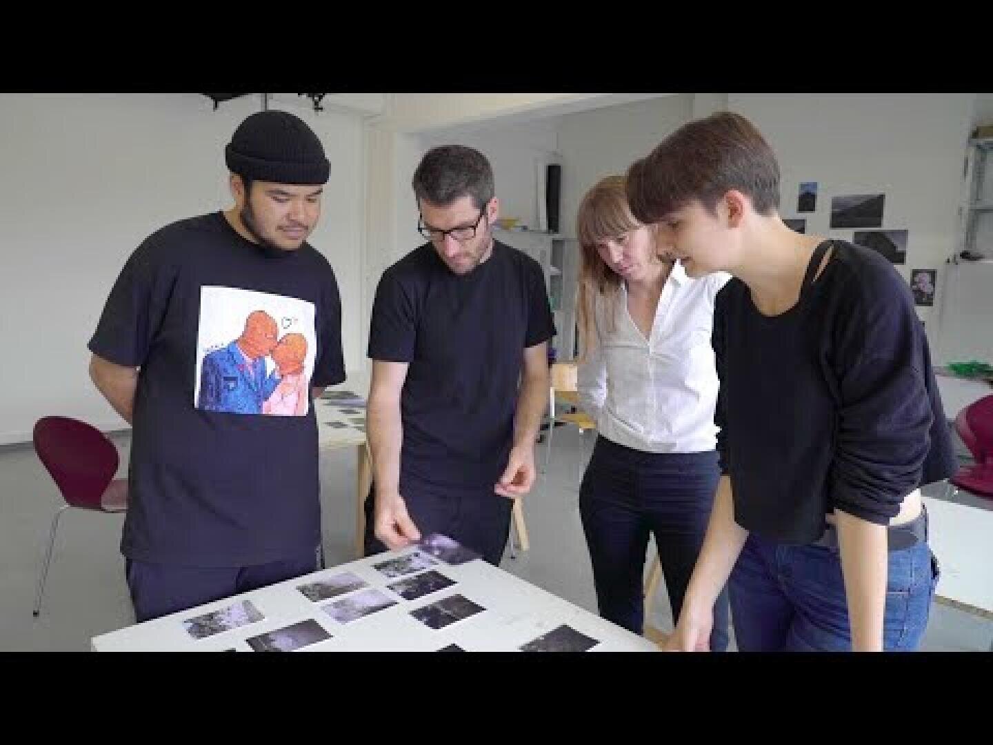 Vier Personen stehen in einem hellen, modernen Raum um einen Tisch herum und betrachten mehrere Fotos, die auf der Tischplatte ausgelegt sind. Sie scheinen die Bilder zu diskutieren oder gemeinsam zu betrachten.