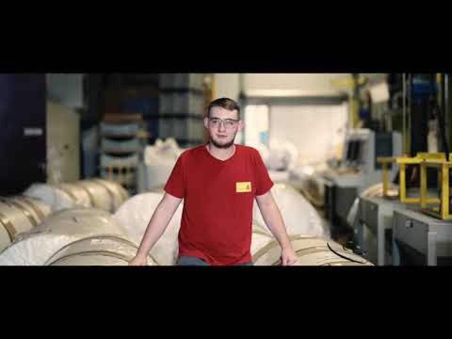 Eine Person mit Brille und rotem T-Shirt steht in einem Industrielager, umgeben von großen Materialrollen und verschiedenen Geräten.