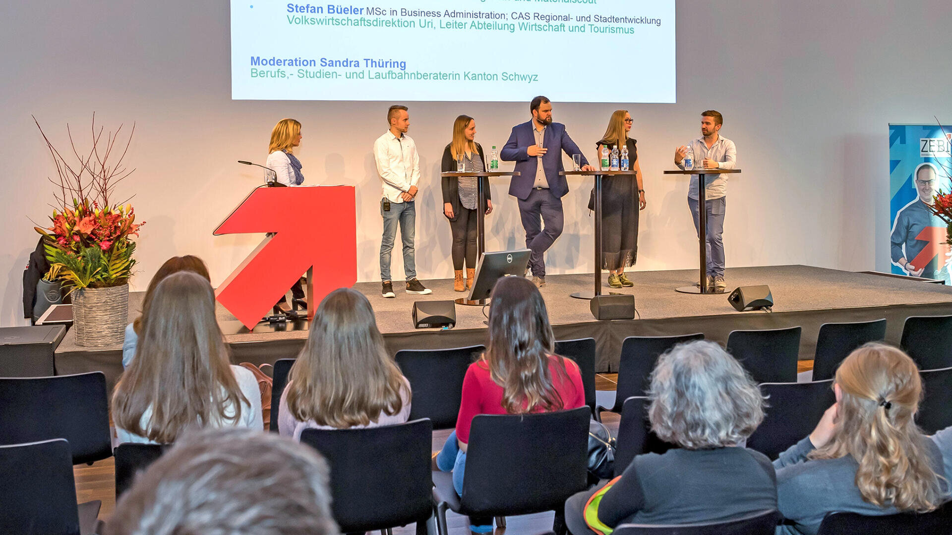 Eine sechsköpfige Gruppe von Personen steht auf der Bühne und diskutiert vor einem Publikum, während hinter ihnen eine Präsentation projiziert wird. Blumen schmücken die linke Seite der Bühne. Das Publikum sitzt in Reihen mit Blick auf die Redner.