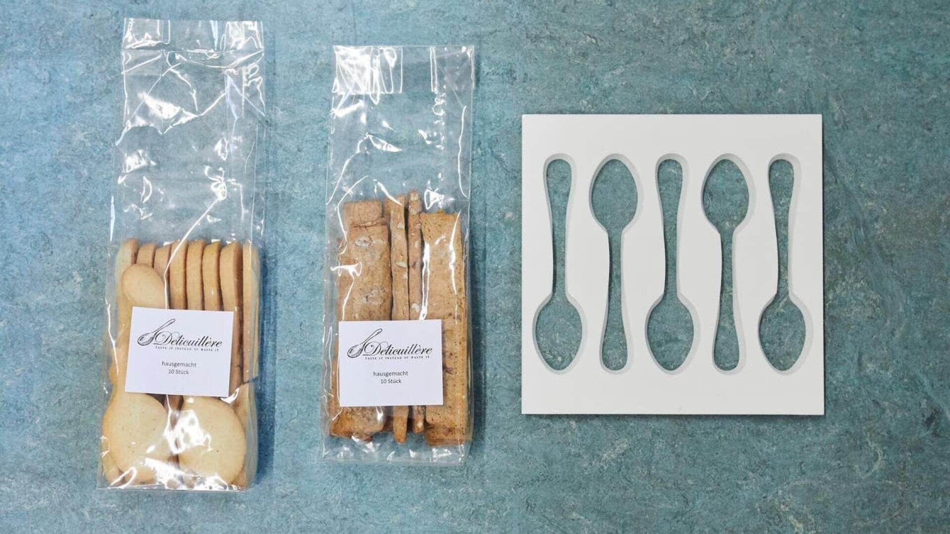Zwei durchsichtige Tüten mit Keksen, eine mit runden Keksen und eine mit rechteckigen Biscotti, werden neben einer weißen Schablone mit vier löffelförmigen Ausschnitten auf einer blauen Fläche platziert.