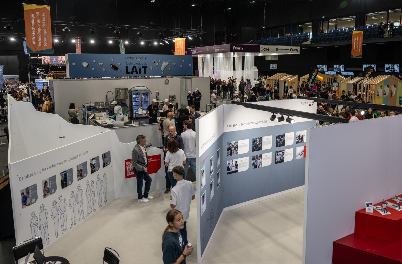 Eine belebte überdachte Ausstellungshalle, in der die Menschen zwischen weißen Ausstellungsständen und Informationstafeln mit verschiedenen Ständen, Bannern und interaktiven Bereichen umhergehen. Die Atmosphäre ist lebendig und einnehmend.