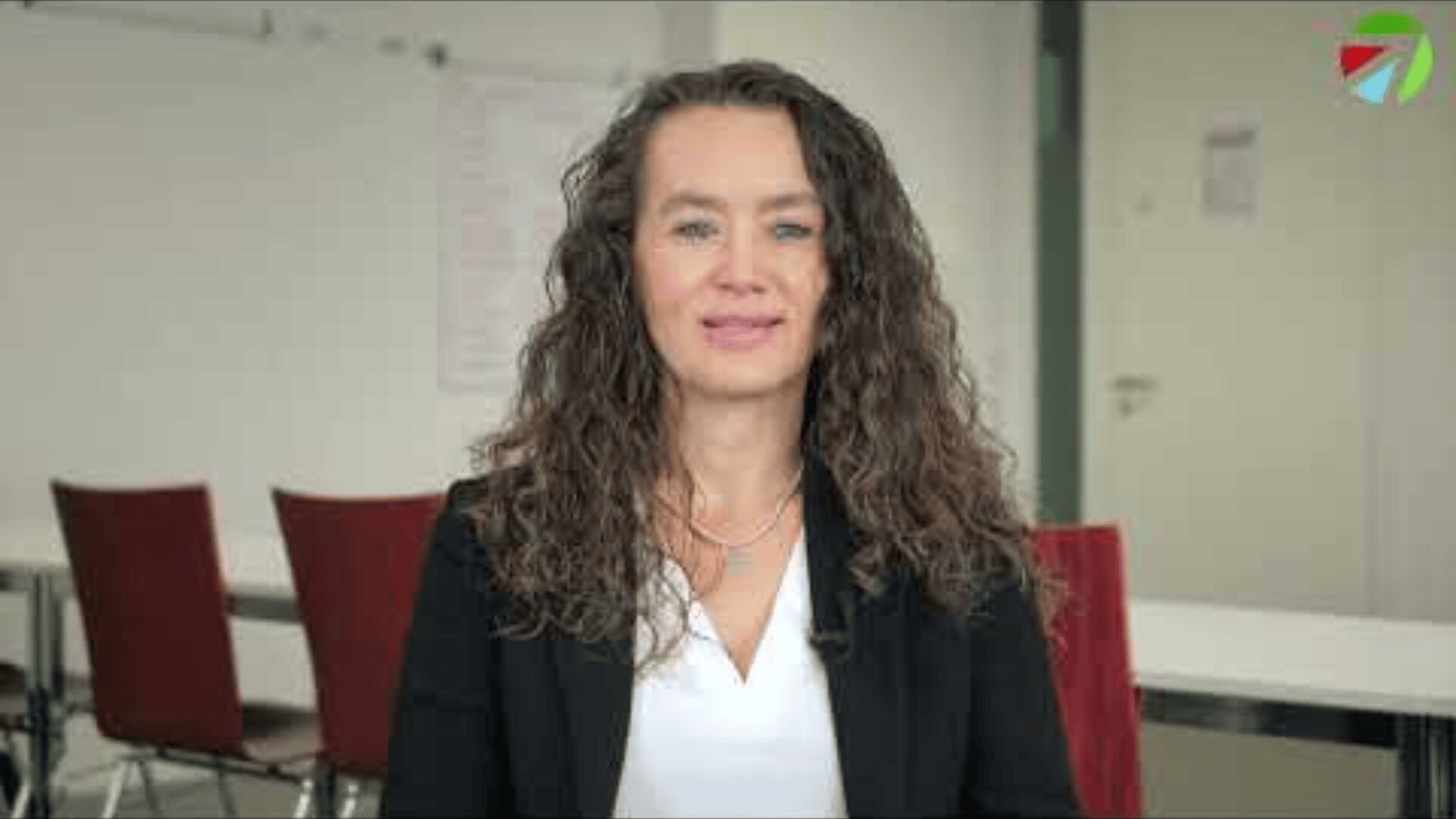 Eine Frau mit langen lockigen braunen Haaren, die einen schwarzen Blazer und eine weiße Bluse trägt, sitzt in einem modernen Büro mit roten Stühlen und einem Whiteboard hinter ihr.