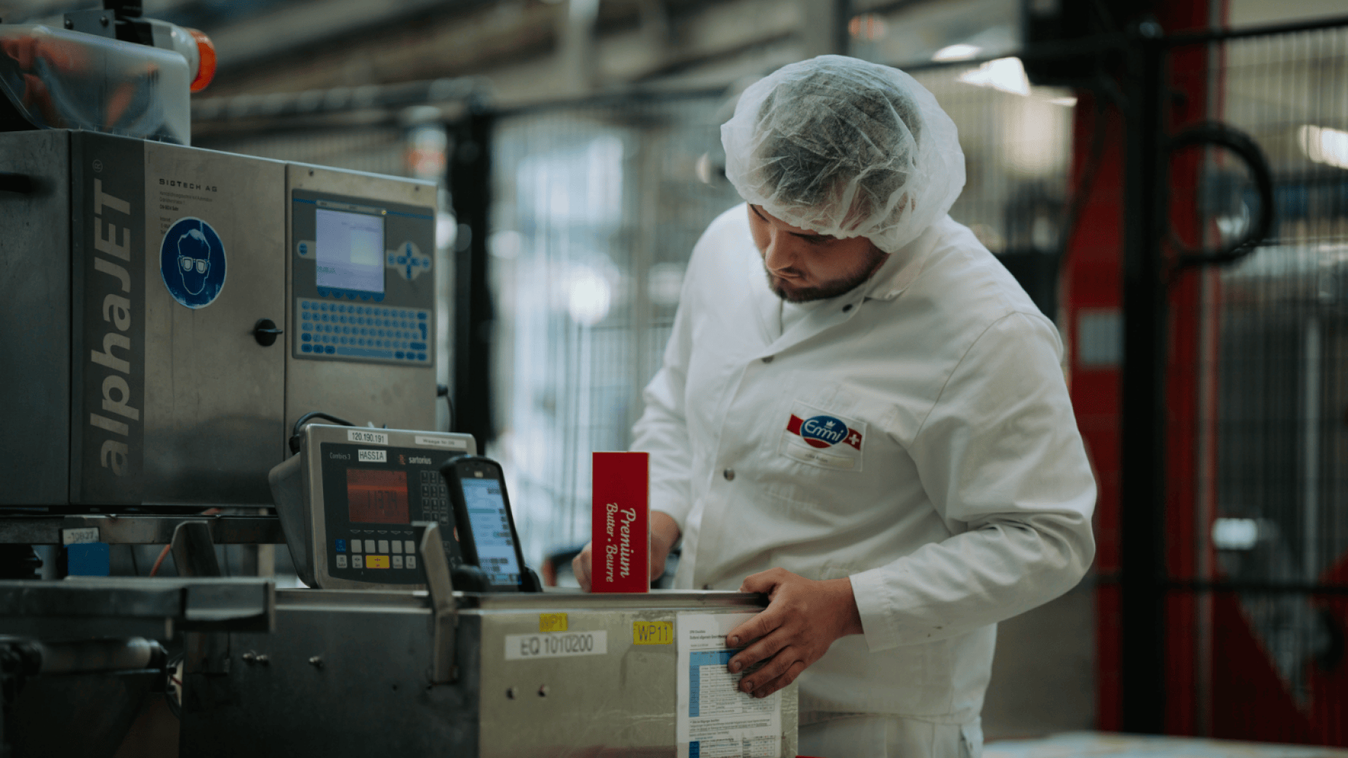 Ein Fabrikarbeiter in weißem Kittel und Haarnetz überprüft an einer Industriemaschine in einer Produktionsstätte die Verpackungen.