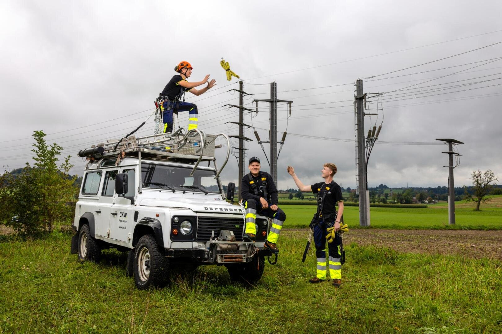 Drei Mitarbeiter eines Energieversorgungsunternehmens in Schutzkleidung und mit Sicherheitsgurten sind auf einem Feld in der Nähe von Stromleitungen um ein weißes Servicefahrzeug herum beschäftigt.