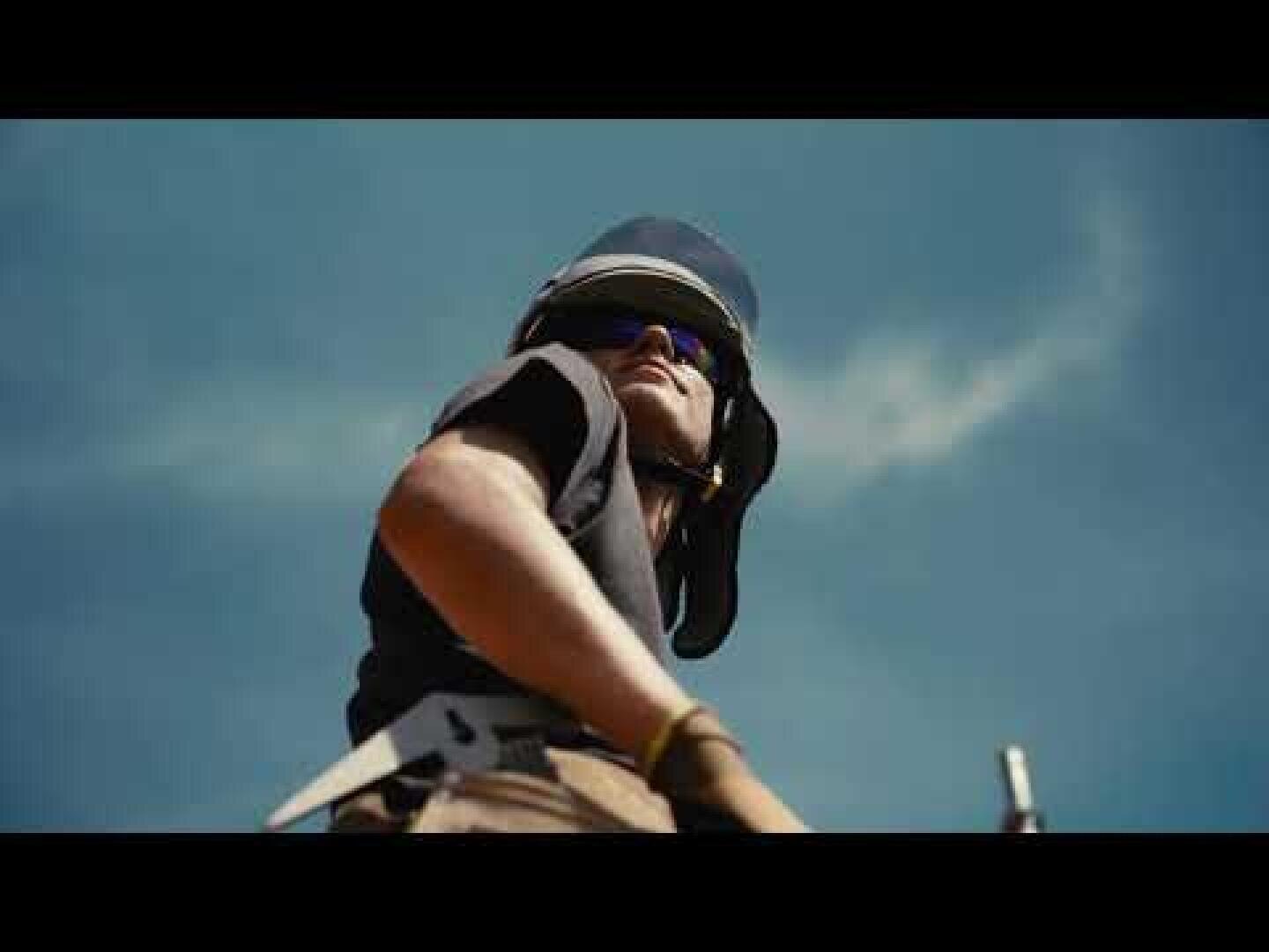 Eine Person mit Helm und Sonnenbrille steht im Freien und blickt in den Himmel, im Hintergrund sind ein blauer Himmel und vereinzelte Wolken zu sehen. Das Foto wurde aus einem niedrigen Blickwinkel aufgenommen.
