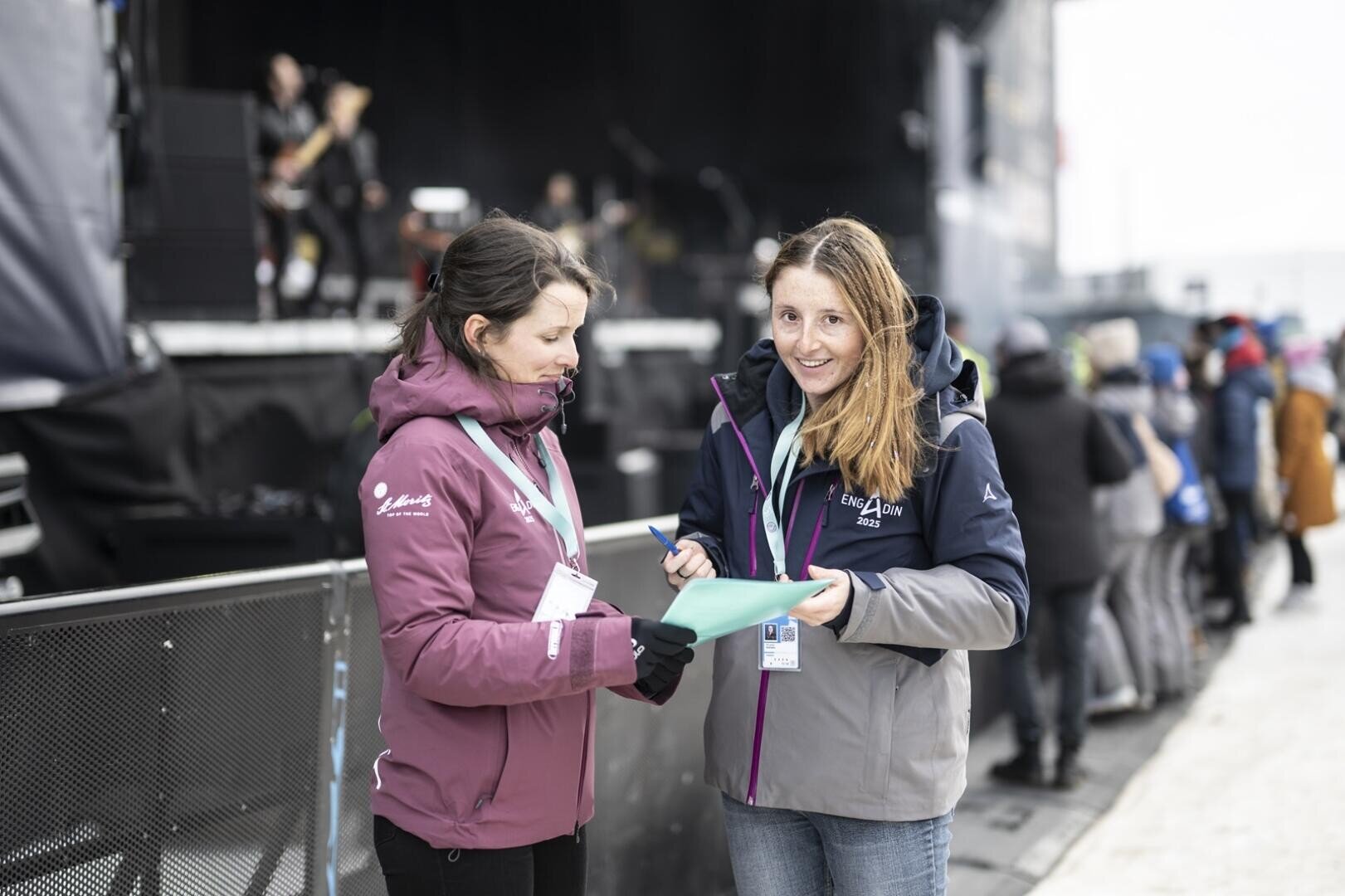 Zwei Frauen in Jacken stehen bei an der Freestyle-WM in der Nähe einer Bühne. Im Hintergrund steht eine Menschenmenge mit Blick auf die Bühne, auf der eine Band auftritt. Beide Frauen tragen Veranstaltungsausweise.