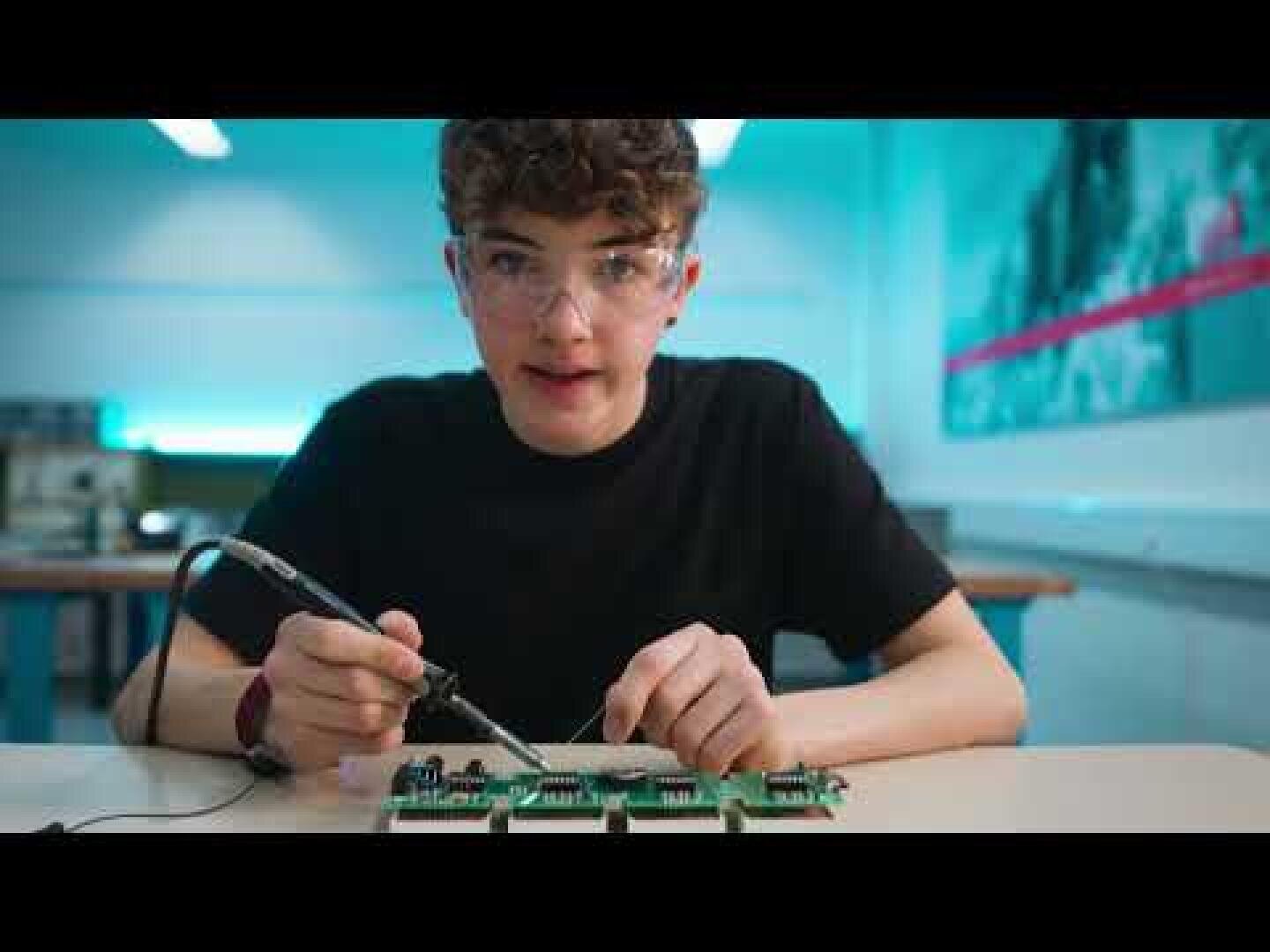 Ein Jugendlicher mit Schutzbrille arbeitet mit einem Lötkolben an einer Leiterplatte in einem hell erleuchteten Klassenzimmer oder Labor.