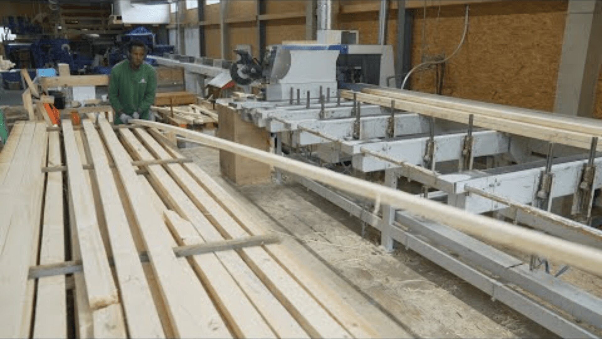 Ein Arbeiter in einem grünen Hemd bedient die Maschinen in einer Holzverarbeitungsfabrik. Er hantiert mit langen Holzbrettern an einem Fließband, das von Holzstapeln umgeben ist.