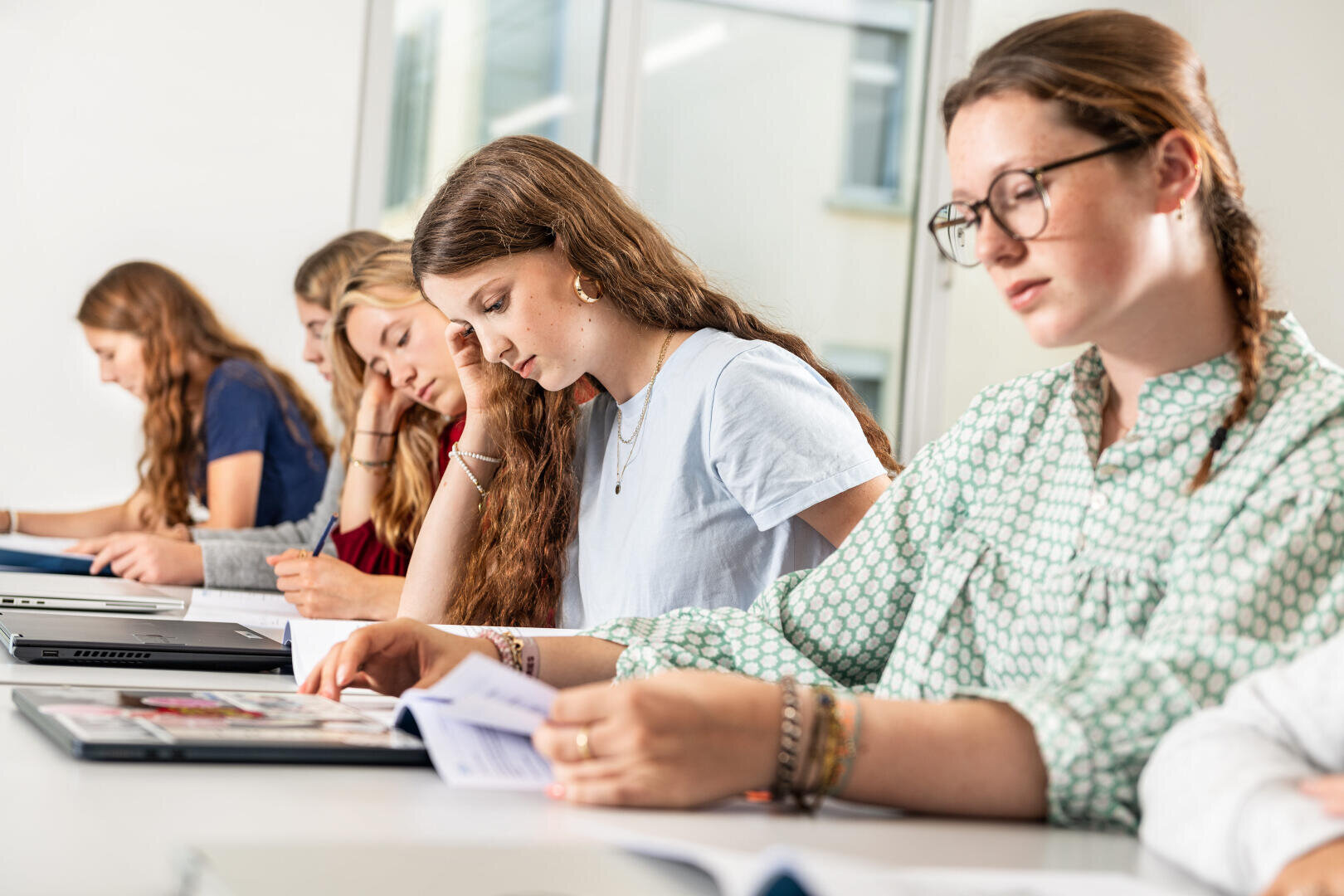 Fünf junge Frauen sitzen in einem hellen Klassenzimmer in einer Reihe an ihren Schreibtischen und sind ganz auf das Lesen und die Arbeit mit Unterlagen und Laptops konzentriert.
