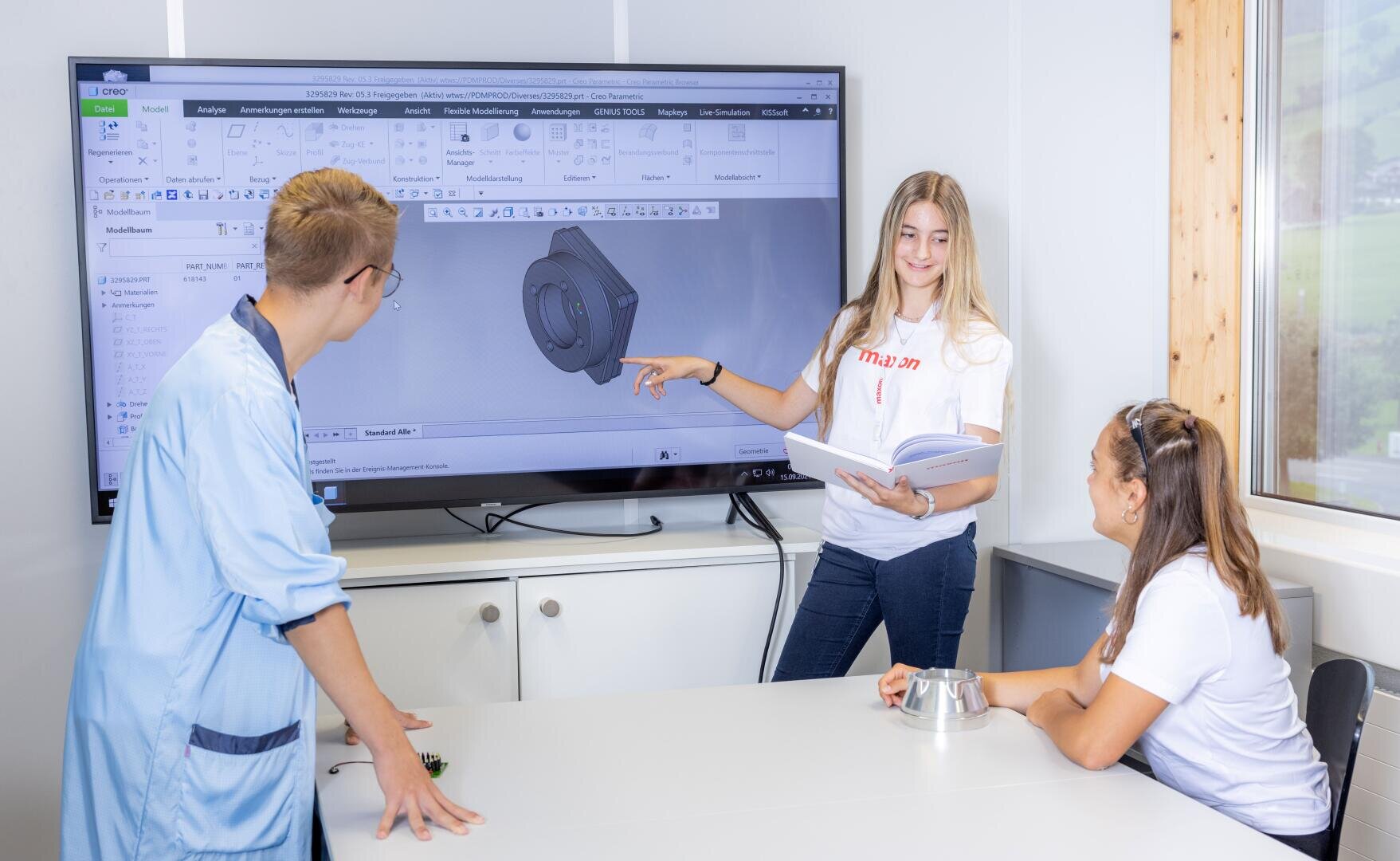 Drei junge Leute diskutieren in einem hellen Raum über ein mechanisches 3D-Modell, das auf einem großen Bildschirm angezeigt wird. Eine Person zeigt auf den Bildschirm, während die anderen zusehen und sich Notizen machen. Vor ihnen auf dem Tisch liegt ein Metallobjekt.