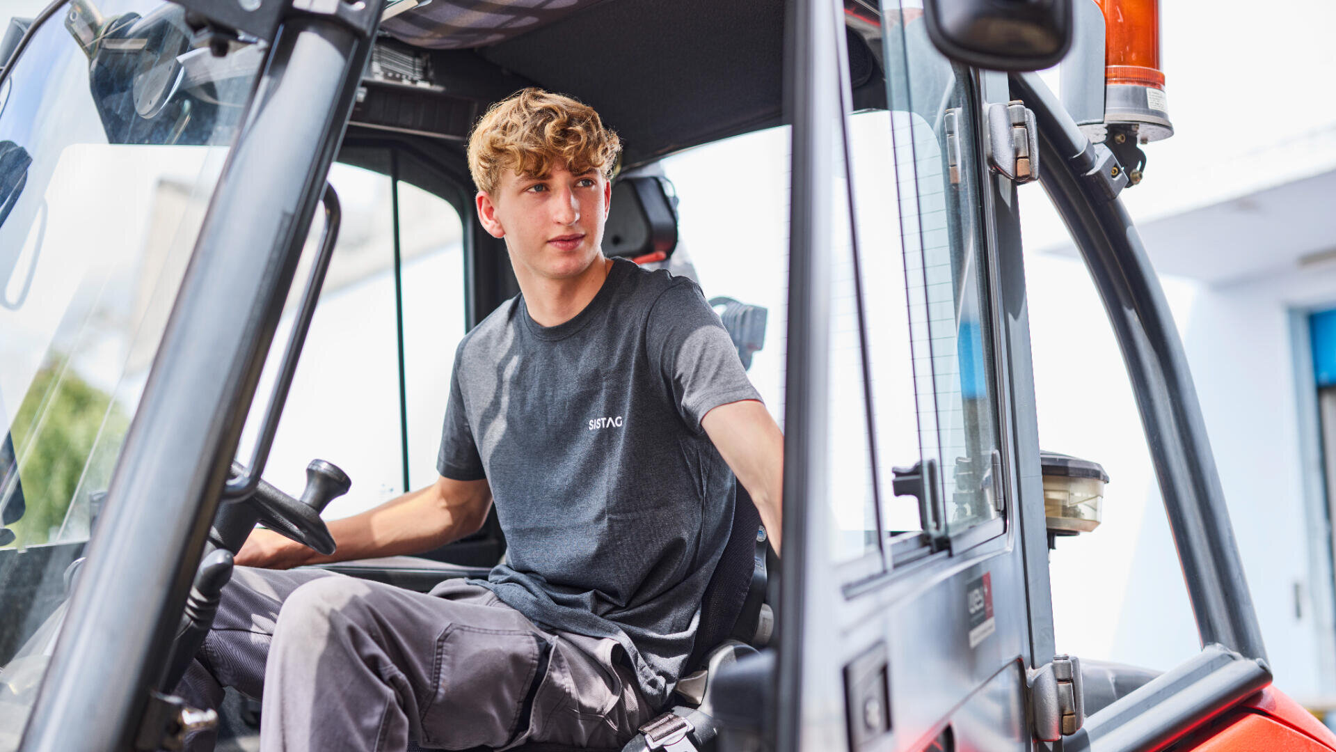 Ein junger Mann mit lockigem Haar sitzt auf dem Fahrersitz eines Fahrzeugs, möglicherweise eines Traktors, und schaut aus der offenen Tür zu seiner Linken, während eine Hand auf dem Lenkrad ruht.