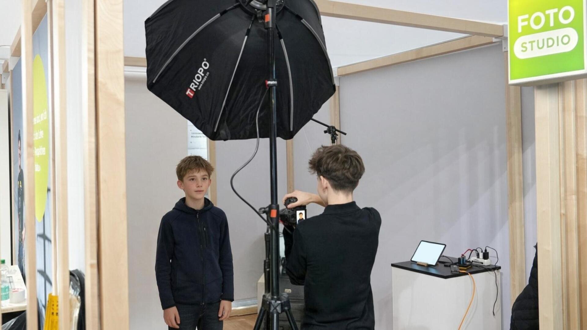 Zwei Jungen in einem kleinen Fotostudio; einer steht als Motiv da, während der andere mit einer Kamera und einem Stativ ein Foto macht. Außerdem sind in der Szene eine große Softbox und ein kleiner Ausrüstungstisch zu sehen.