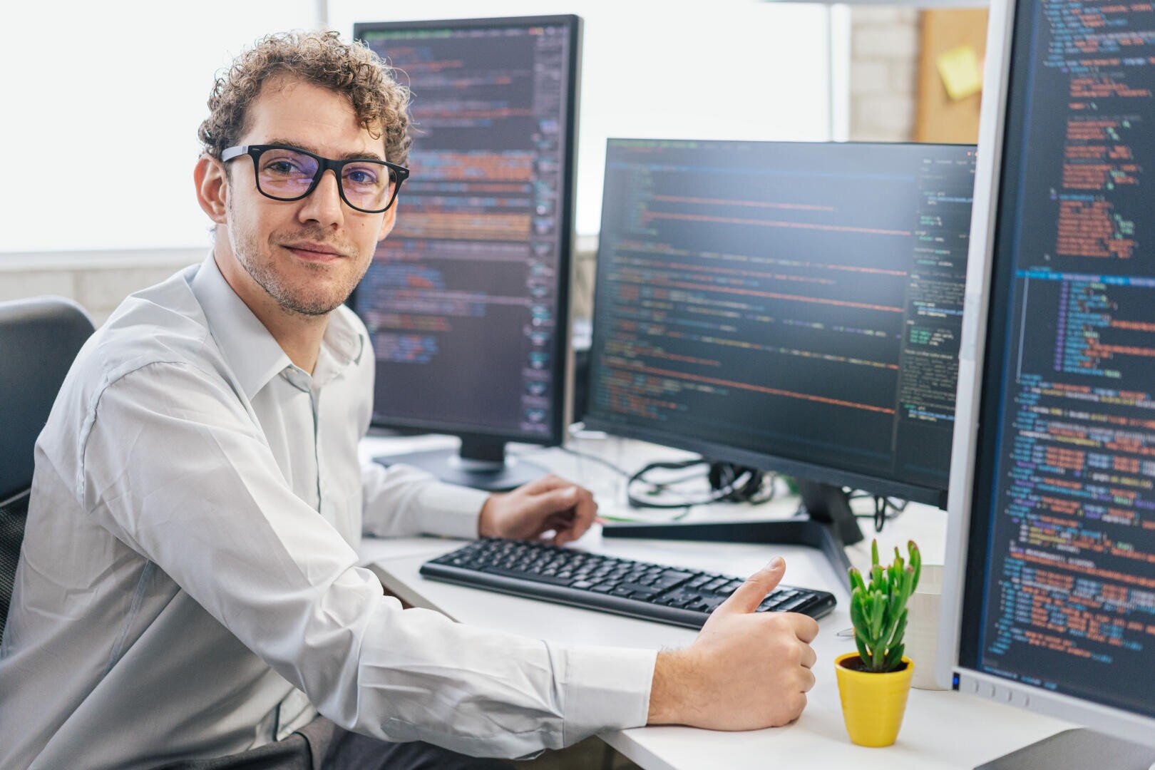 Ein Mann mit Brille und weißem Hemd sitzt an einem Schreibtisch mit drei Monitoren, auf denen Codes angezeigt werden. Er schaut in die Kamera und zeigt den Daumen nach oben. Eine kleine Topfpflanze steht neben der Tastatur.