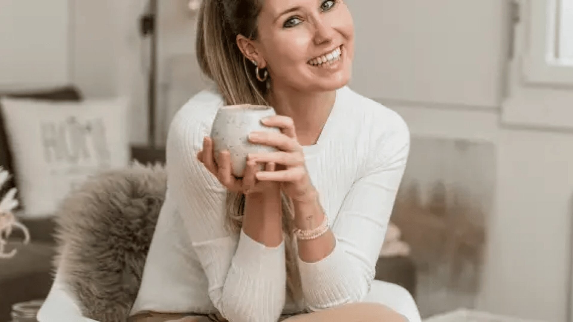 Eine Frau mit zurückgebundenen langen Haaren lächelt, während sie drinnen sitzt und mit beiden Händen einen Keramikbecher hält. Sie trägt einen weißen Pullover und eine hellbraune Hose, im Hintergrund ist eine gemütliche Einrichtung zu sehen.