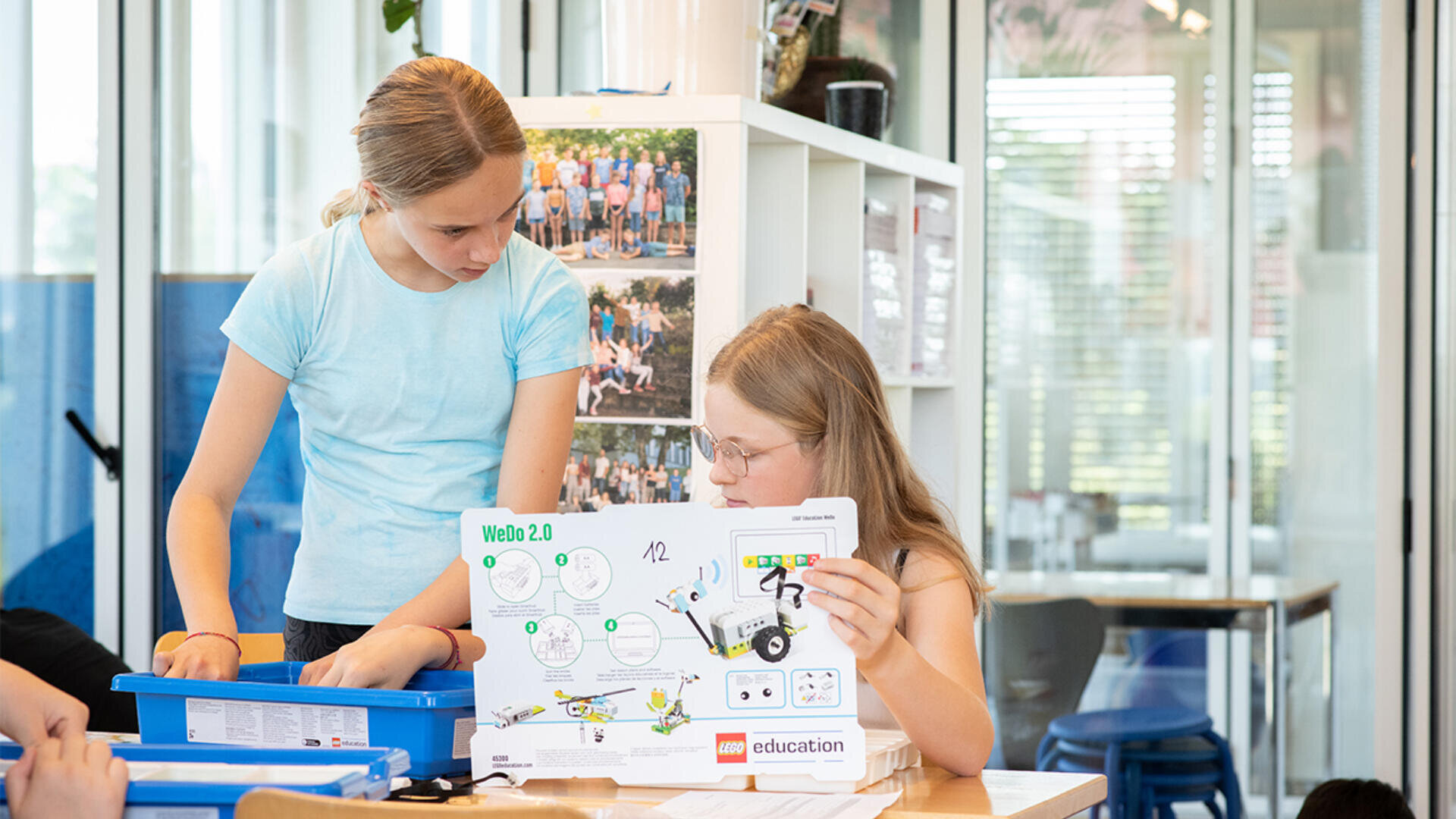 Zwei Mädchen arbeiten gemeinsam an einem Tisch an einem LEGO-Roboterprojekt. Ein Mädchen hält eine Bedienungsanleitung in der Hand und liest sie, während das andere in einen blauen Behälter schaut, beide konzentriert und engagiert in einem hellen Klassenzimmer.