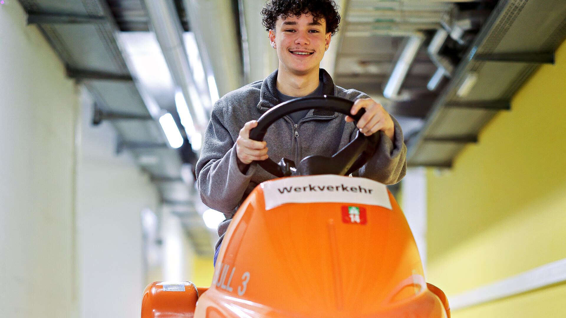 Ein lächelnder junger Mann mit lockigem Haar fährt ein orangefarbenes Fahrzeug mit der Aufschrift Werkverkehr durch einen hellen Innenkorridor mit freiliegenden Rohren und Metallträgern an der Decke.