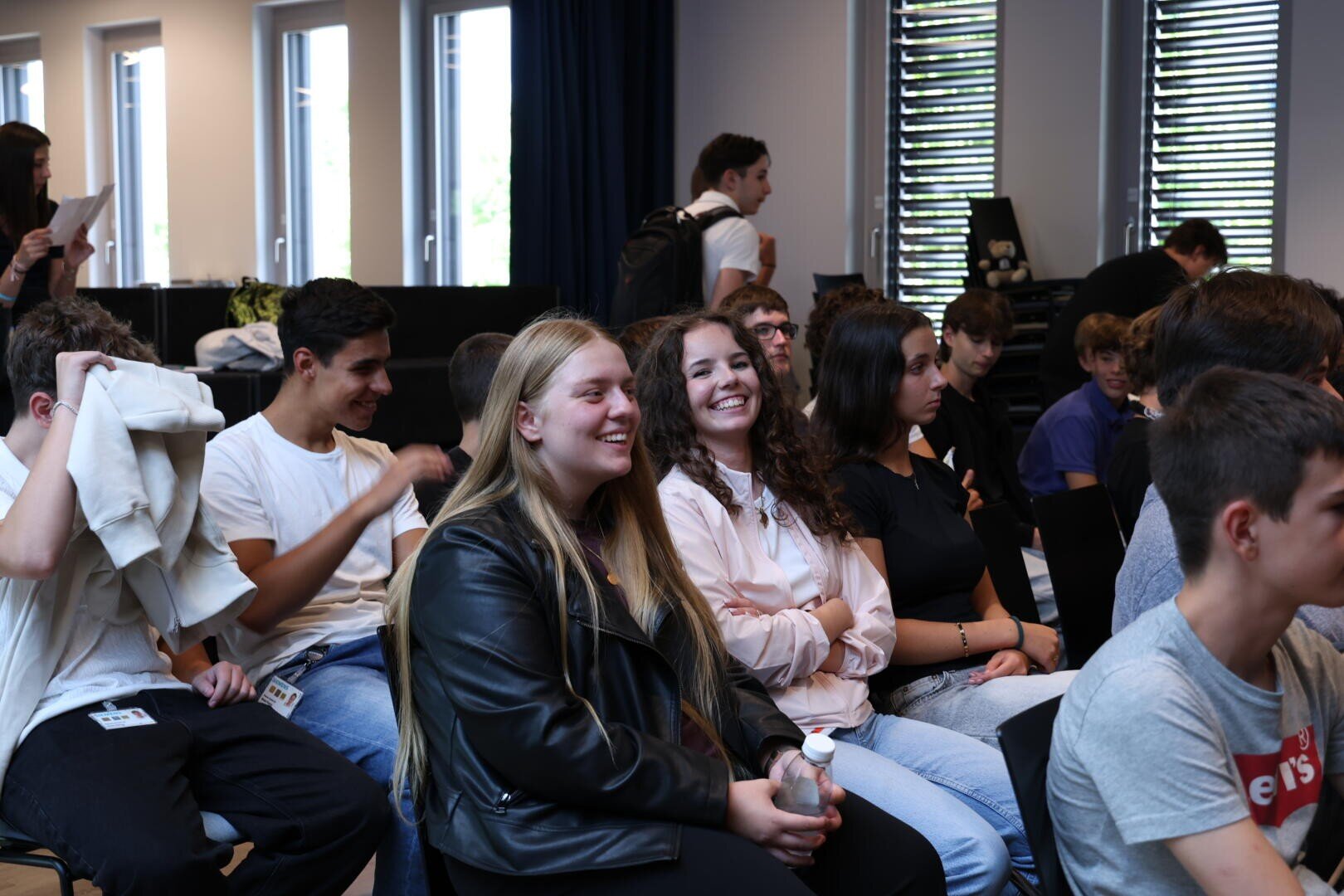 Eine Gruppe Jugendlicher sitzt in einem hell erleuchteten Klassenzimmer zusammen, einige lächeln und unterhalten sich, während andere aufmerksam nach vorne schauen. Die Atmosphäre wirkt lebendig und freundlich.