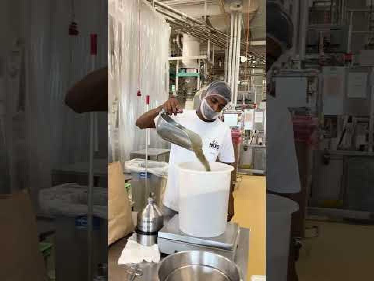 Eine Person mit Haarnetz und Gesichtsmaske gießt Flüssigkeit aus einem Metallkrug in einen großen weißen Behälter in einer Fabrik- oder Laborumgebung mit Industrieanlagen und Rohren im Hintergrund.