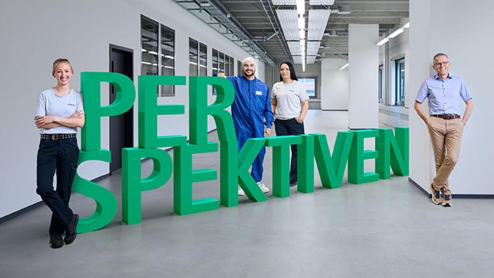 Vier Personen stehen in einem hellen, modernen Flur neben großen grünen 3D-Buchstaben mit dem Wort „PERSPEKTIVEN“. Sie lächeln und posieren lässig und stehen für Vielfalt und Teamwork.