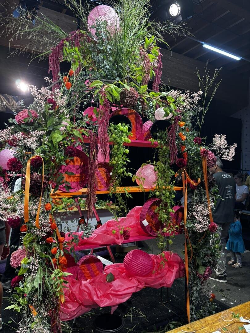 Ein farbenfroher Blumenbogen, geschmückt mit rosa Stoff, Laternen, üppigem Grün und herabhängenden Blumen, steht in einem Innenraum unter heller Beleuchtung, im Hintergrund sind Menschen zu sehen.