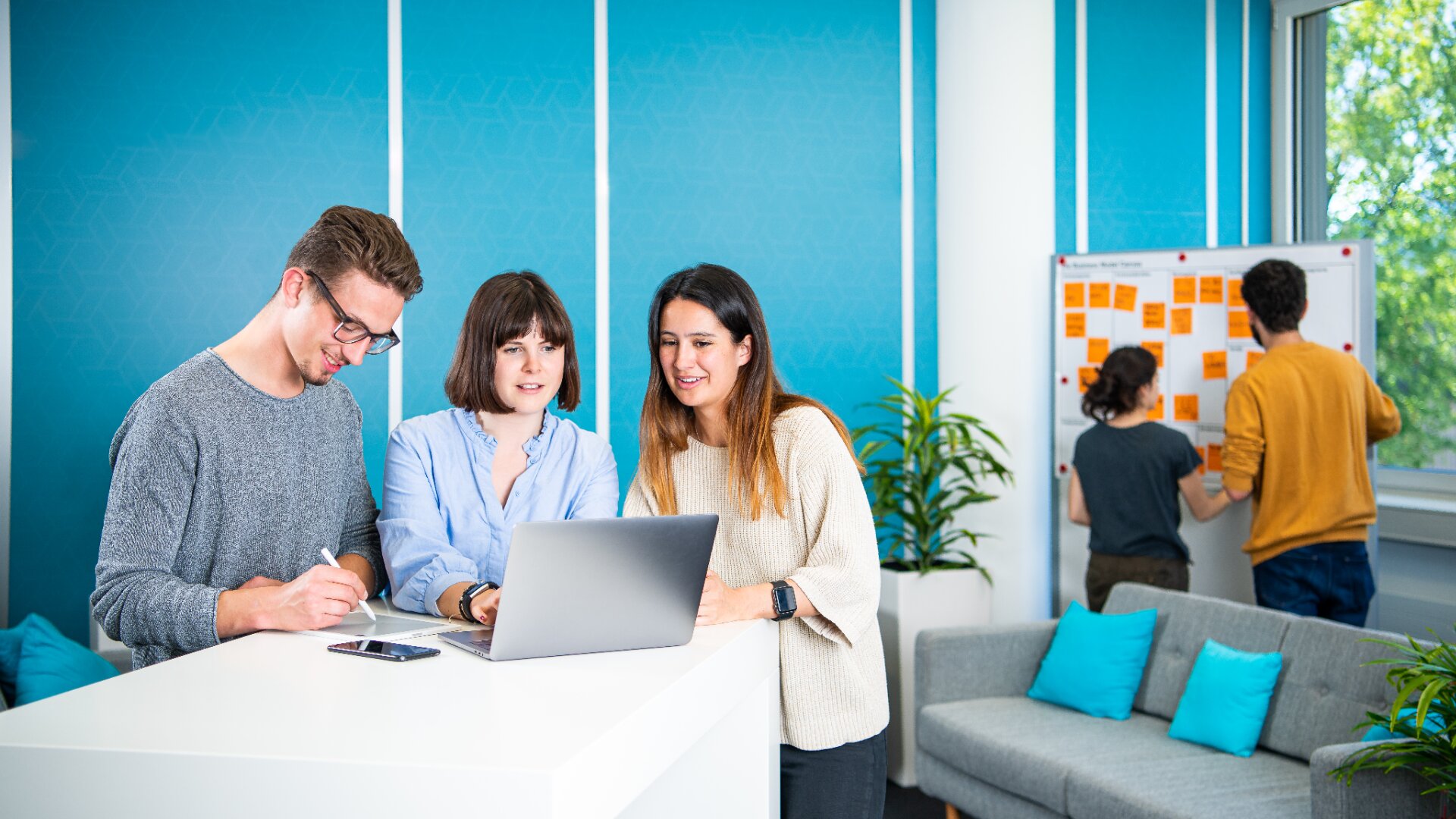 In einem modernen, hellen Büro mit blauen Wänden und großen Fenstern stehen drei Personen an einem weißen Schreibtisch und schauen lächelnd auf einen Laptop, während zwei andere an einem Whiteboard mit Klebezetteln arbeiten.