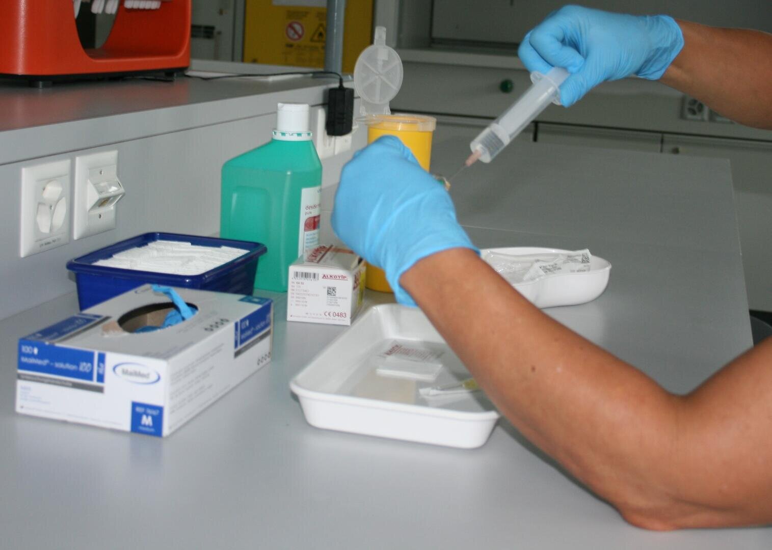 Eine Person mit blauen Handschuhen bereitet eine Spritze an einem medizinischen Arbeitsplatz vor, an dem verschiedene medizinische Hilfsmittel, darunter Handschuhe, Behälter, Schachteln und eine grüne Flasche auf der Arbeitsplatte stehen.