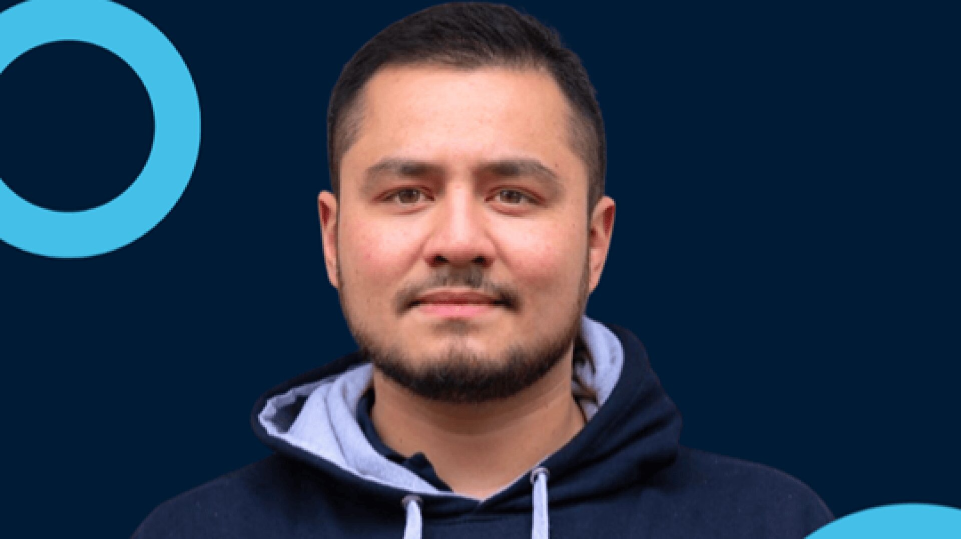 Ein Mann mit kurzen dunklen Haaren und gestutztem Bart, der einen dunklen Kapuzenpulli mit hellgrauer Kapuze trägt, steht vor einem dunkelblauen Hintergrund mit hellblauen Kreisformen.