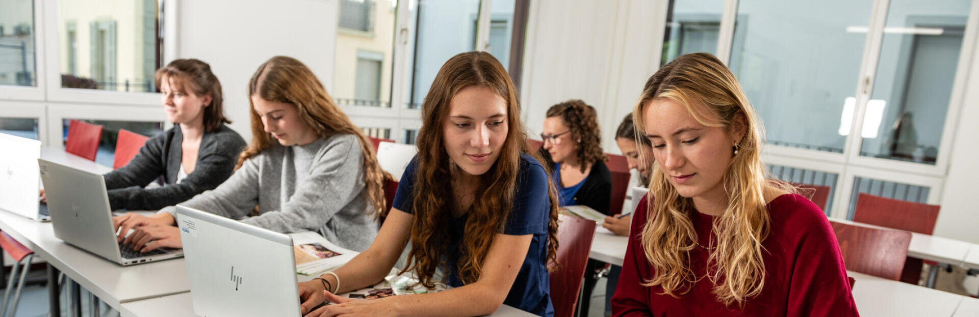 Fünf junge Frauen sitzen an Schreibtischen in einem hellen Klassenzimmer, arbeiten an ihren Laptops und lesen Unterlagen. Einige sind ganz auf ihre Bildschirme konzentriert, während andere sich miteinander unterhalten.